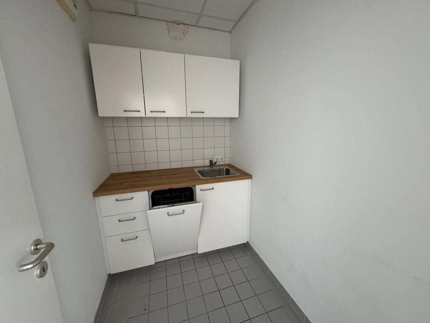 Kleine Küchenzeile mit weißen Schränken, Spüle, Geschirrspüler und Fliesenboden in hellem Grau.