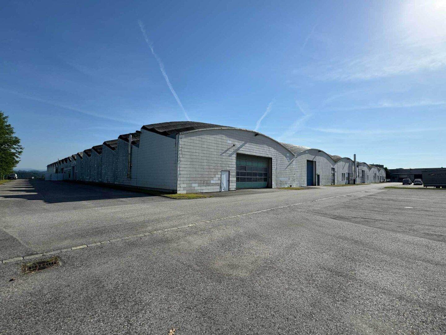 Weitläufige Ansicht eines modernen Industriegebäudekomplexes mit geschwungenen Dächern und großem asphaltierten Vorplatz unter blauem Himmel.