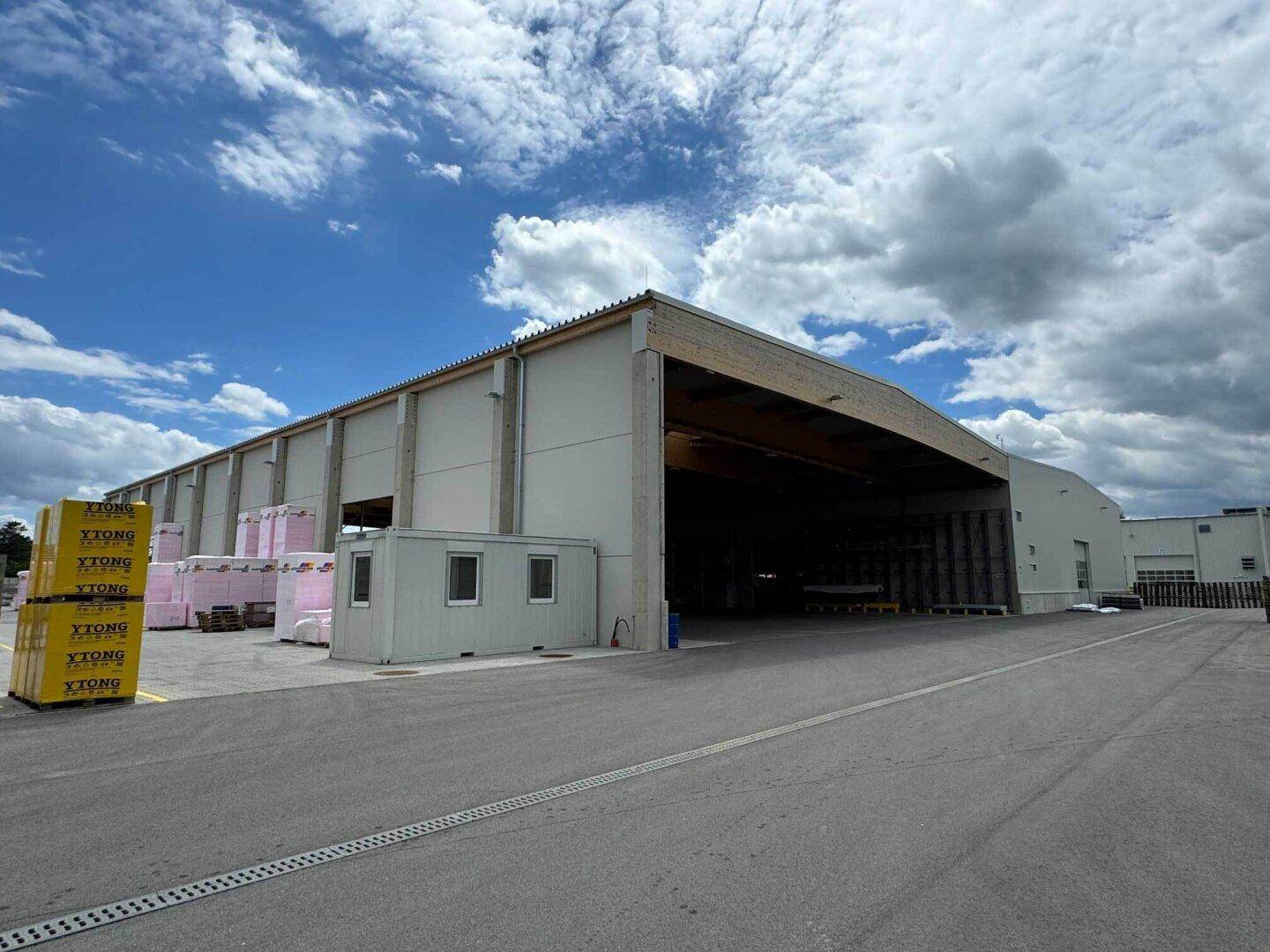 Außenansicht einer modernen Lagerhalle mit offener Ladezone und gestapelten Baumaterialien unter bewölktem Himmel.