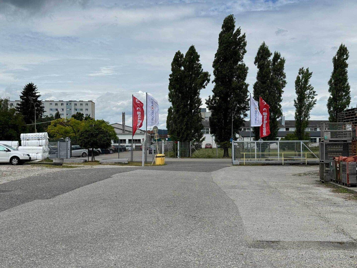 Einfahrt zum Industriegelände mit Fahnen, hohen Bäumen und Blick auf umliegende Gebäude im Hintergrund.