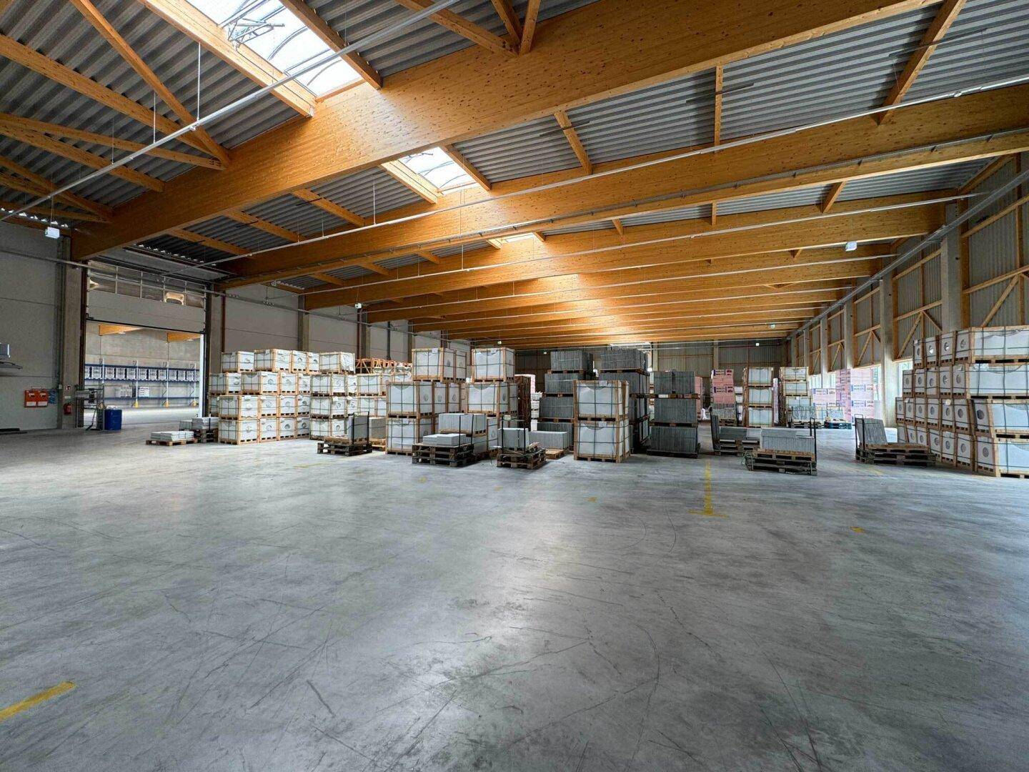 Geräumige Lagerhalle mit hohen Decken, Holzbalken und gestapelten Waren auf Paletten, gut beleuchtet.