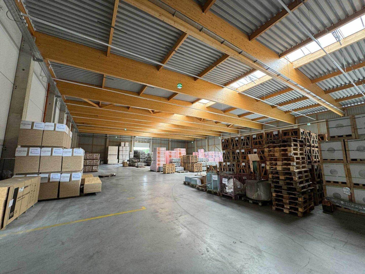 Weitläufige Lagerhalle mit Tageslicht durch Oberlichter und Holzkonstruktion, gefüllt mit Paletten und Waren.