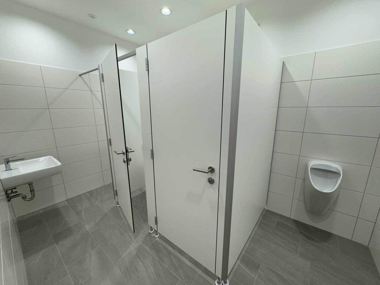 Sauberer, moderner Waschraum mit Urinal, Waschbecken und weißen Trennwänden.