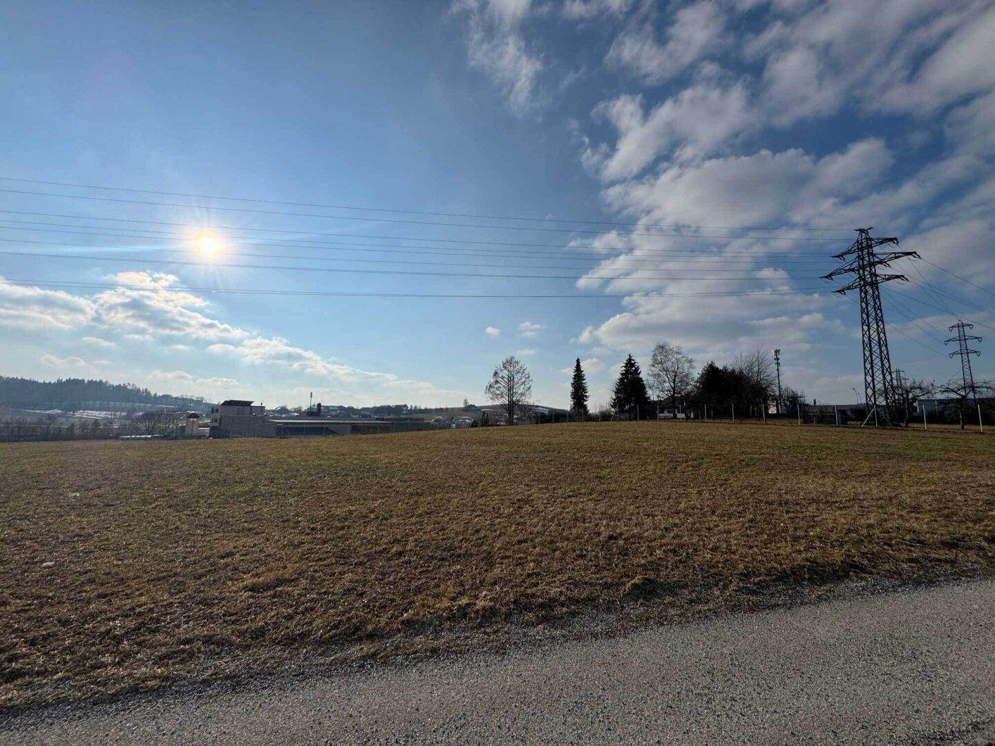 Weitläufiges Feld unter blauem Himmel mit Sonnenschein, im Hintergrund sind Strommasten und Hügel sichtbar.