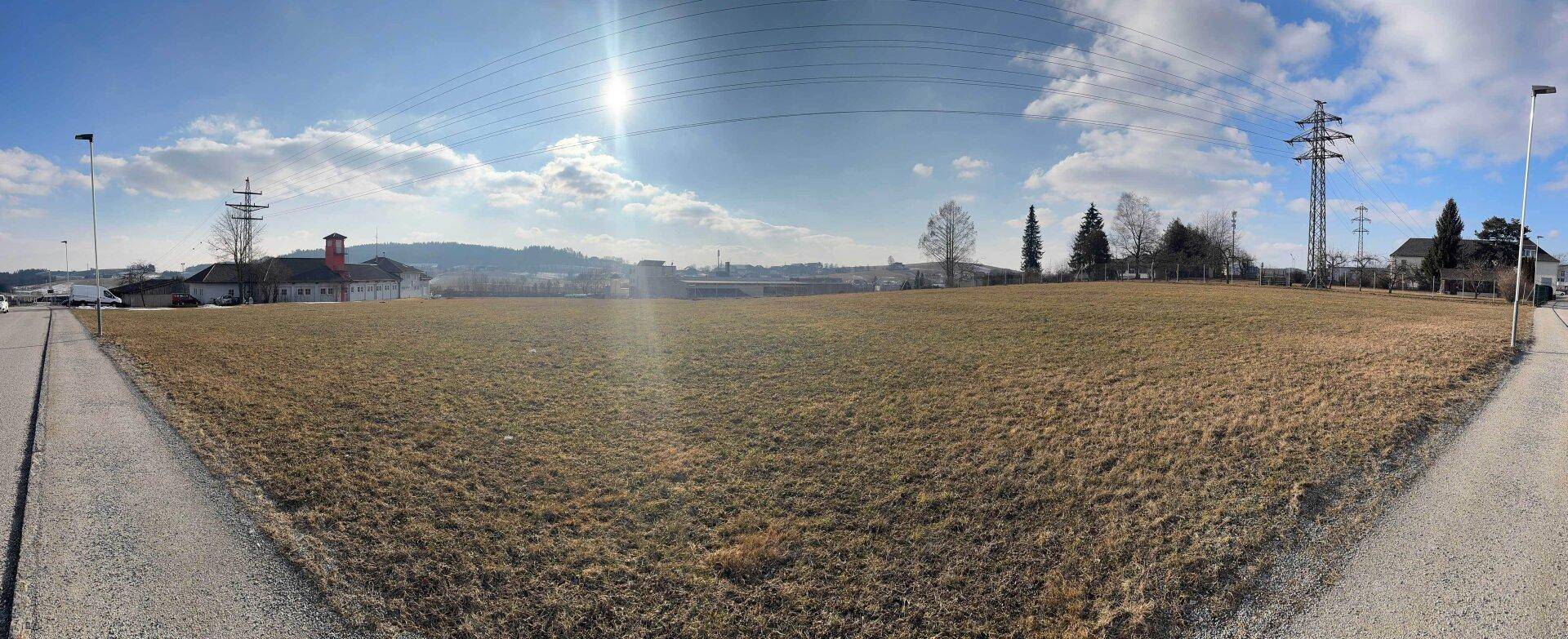 Panoramablick über ein weites Feld mit angrenzender Straße, Strommasten und sonnigem Himmel im Hintergrund.
