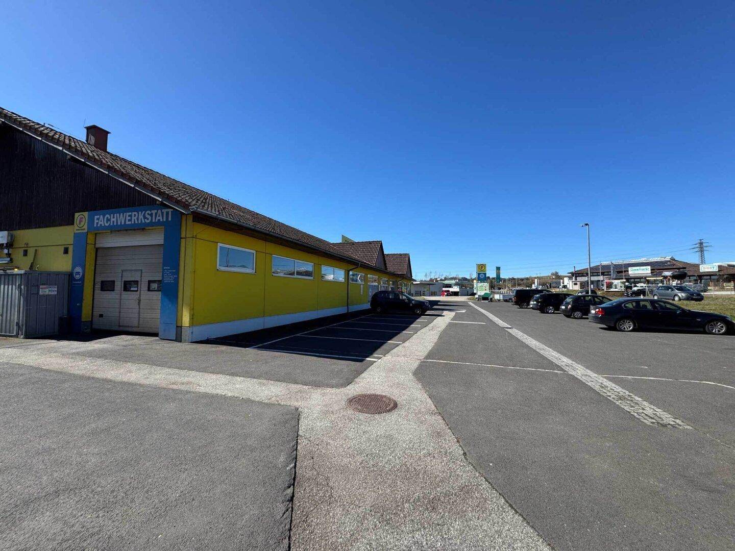 Seitenansicht des Geschäftsgebäudes mit Werkstattbereich und ausgedehntem Parkplatz unter blauem Himmel.