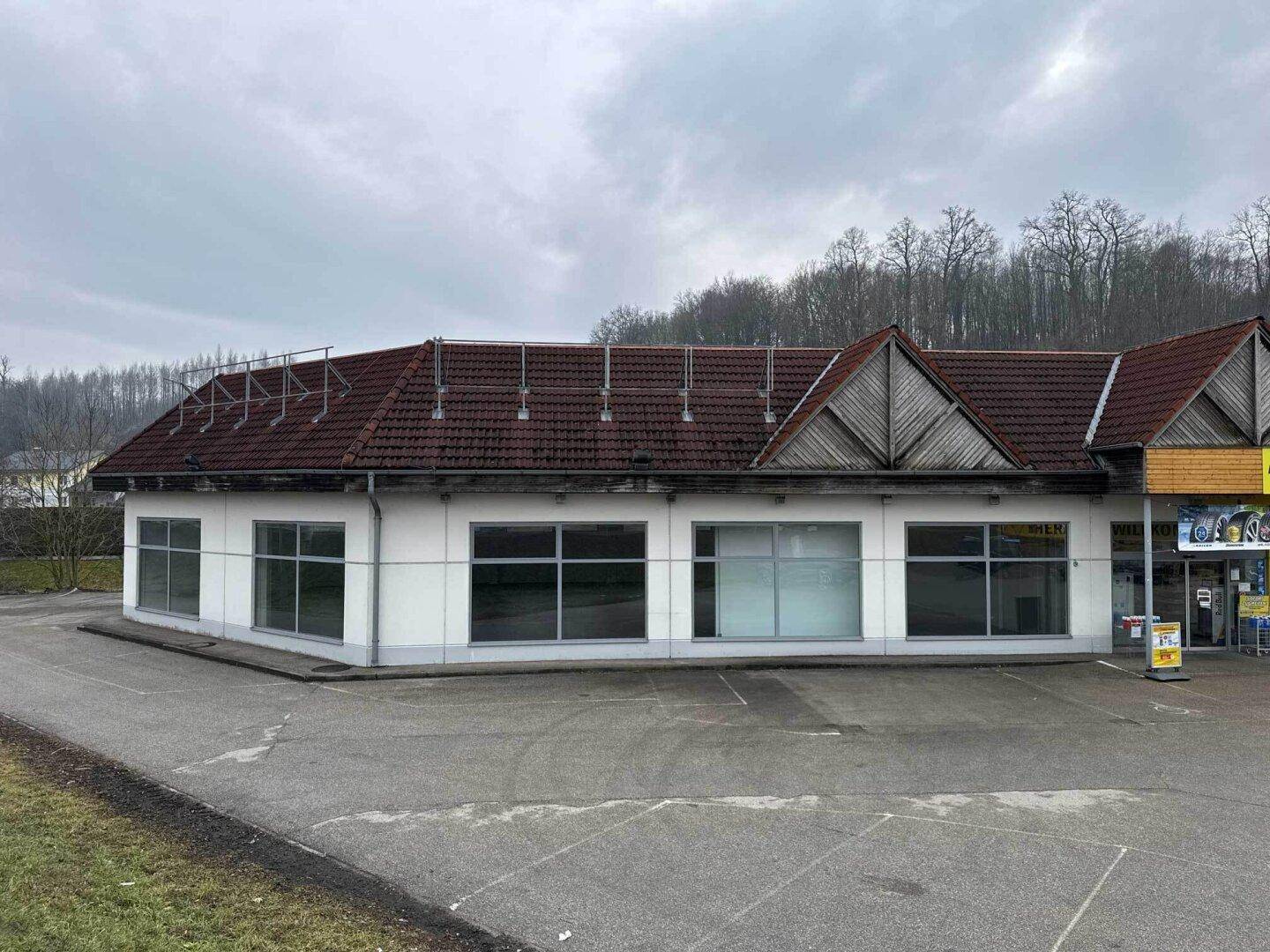 Seitenansicht des Gewerbegebäudes mit großen Fenstern und einem roten Ziegeldach unter bewölktem Himmel.