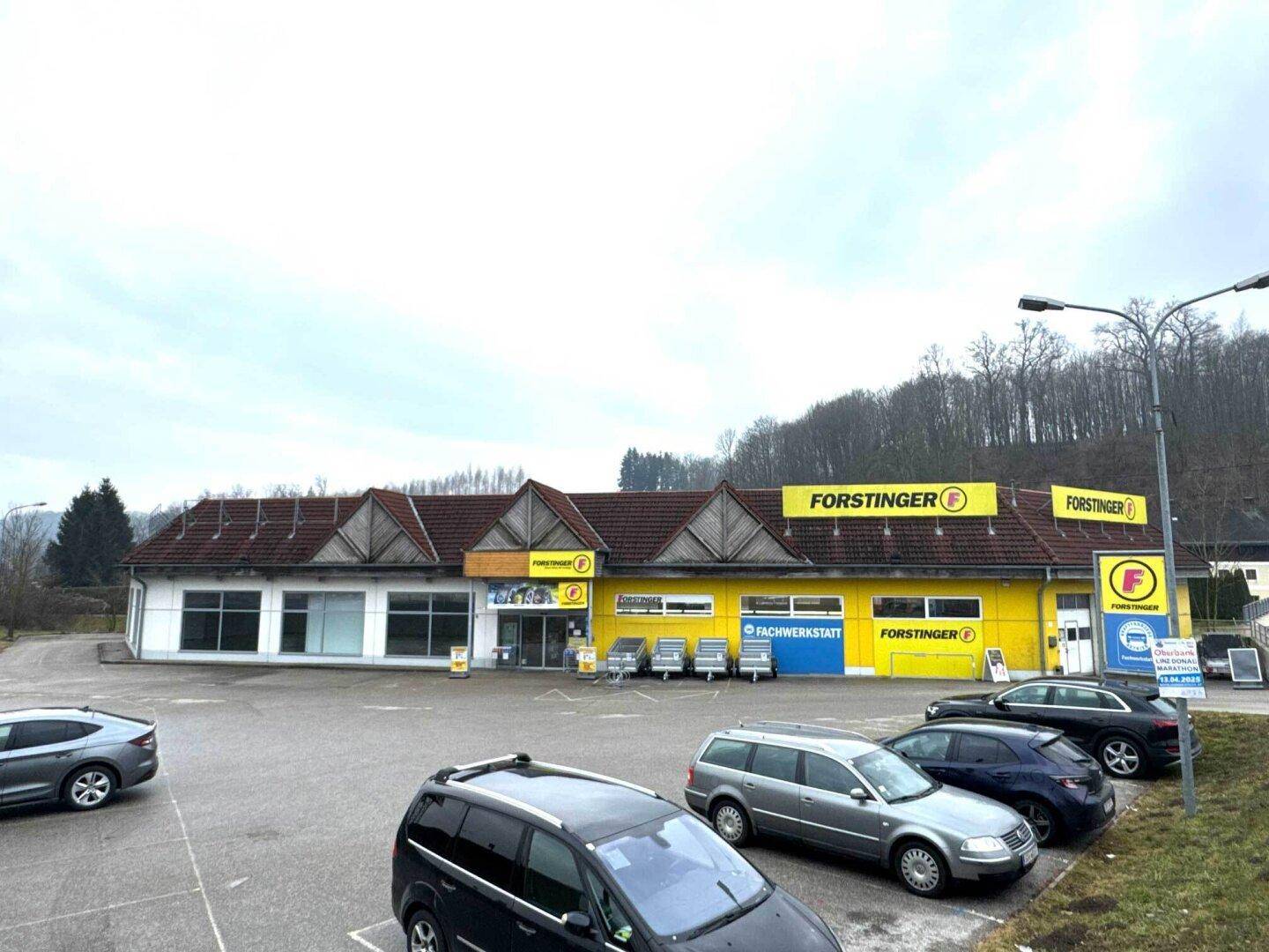 Gewerbegebäude mit gelber Fassade und Parkplätzen im Vordergrund, umgeben von Bäumen und Hügeln.