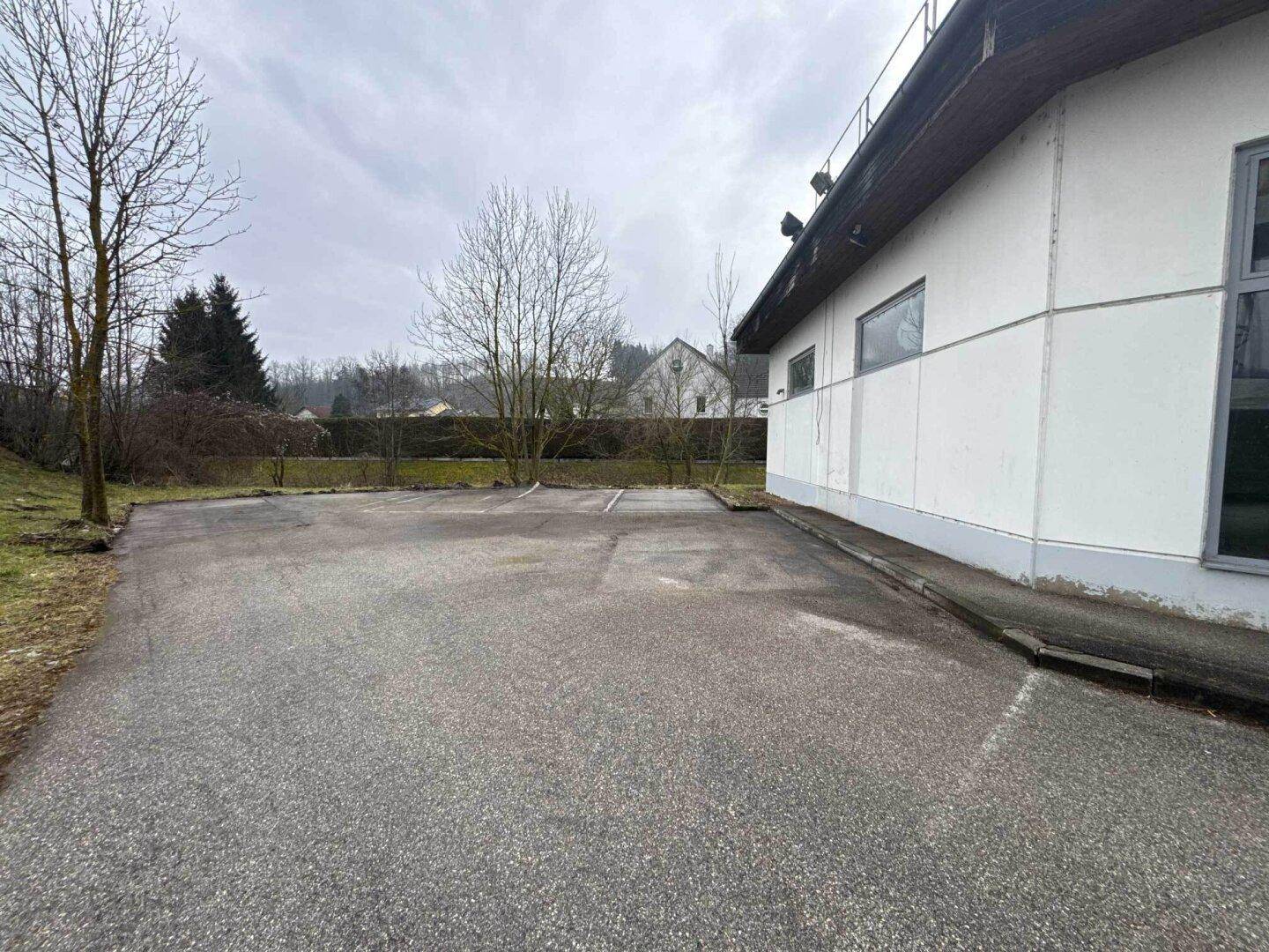Leerer Parkplatz neben dem Gewerbegebäude, umgeben von Bäumen und einer grünen Landschaft.