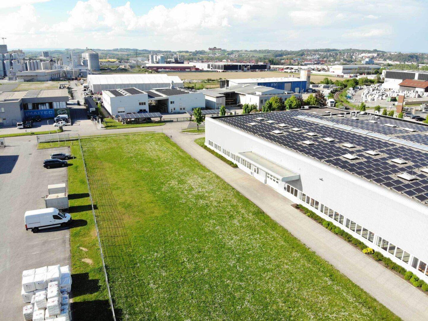 Luftaufnahme eines großen Industriegebäudes mit Solaranlage auf dem Dach und umliegenden Grünflächen.