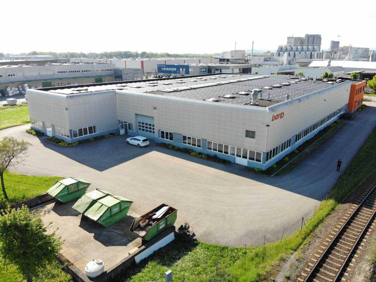 Seitenansicht eines Industriegebäudes mit Laderampe, Parkplatz und Müllcontainern neben Bahngleisen.