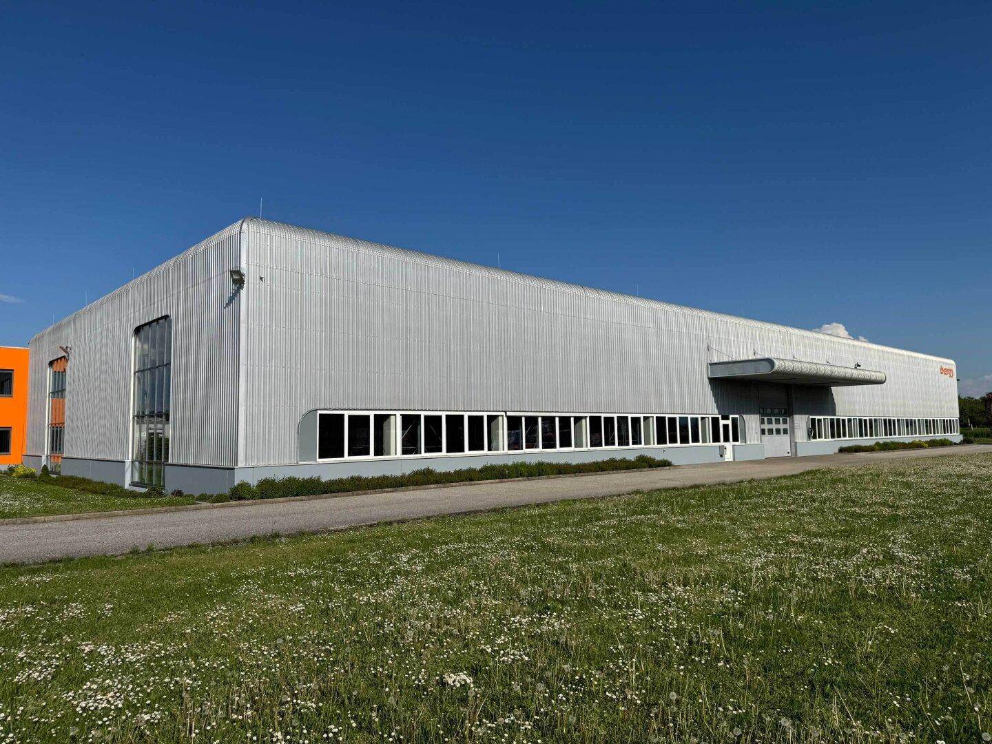 Ansicht des langen Industriegebäudes mit großen Fenstern und umliegender Grünfläche bei Sonnenschein.