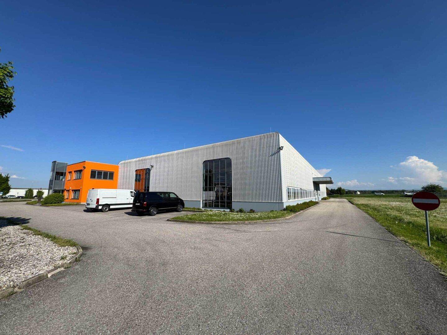 Ansicht des Industriegebäudes mit orangefarbenem Bürotrakt und Zufahrtsstraße unter blauem Himmel.
