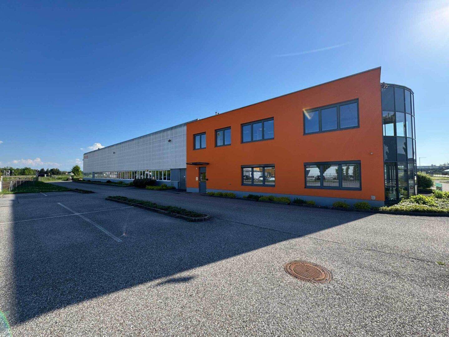 Seitenansicht des orangefarbenen Bürotrakts mit großen Fenstern und angrenzendem Parkplatz.