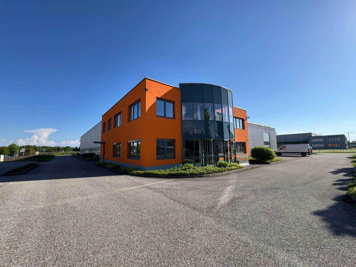 Vorderansicht des orangefarbenen Bürotrakts mit moderner Glasfassade und gepflegter Bepflanzung.
