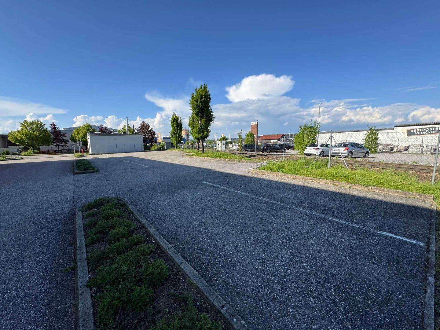 Asphaltierter Parkplatz mit Bäumen und angrenzenden Gebäuden unter einem bewölkten Himmel.