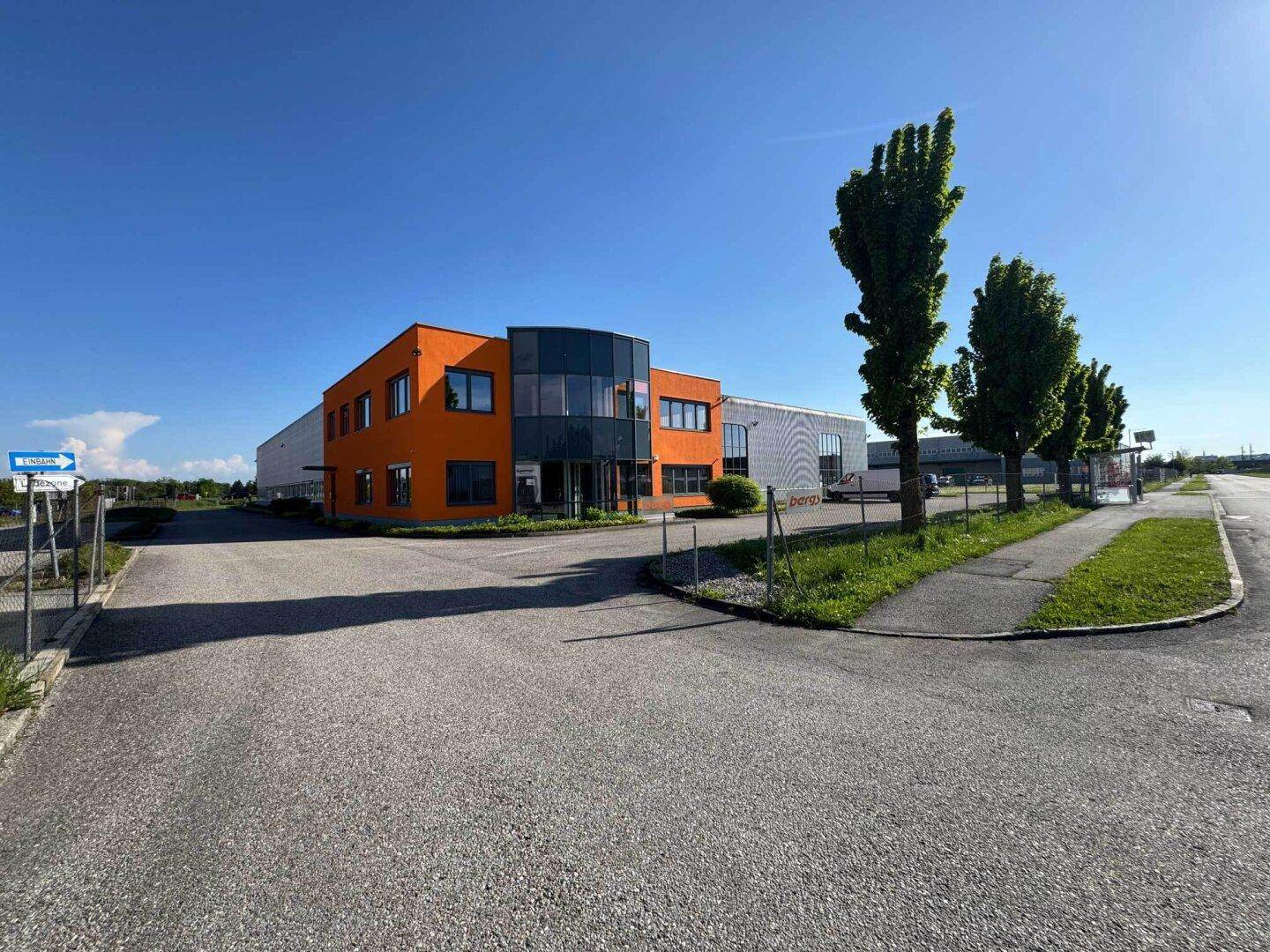 Eingangsbereich des orangefarbenen Bürotrakts mit Glasfassade und Zufahrt unter blauem Himmel.