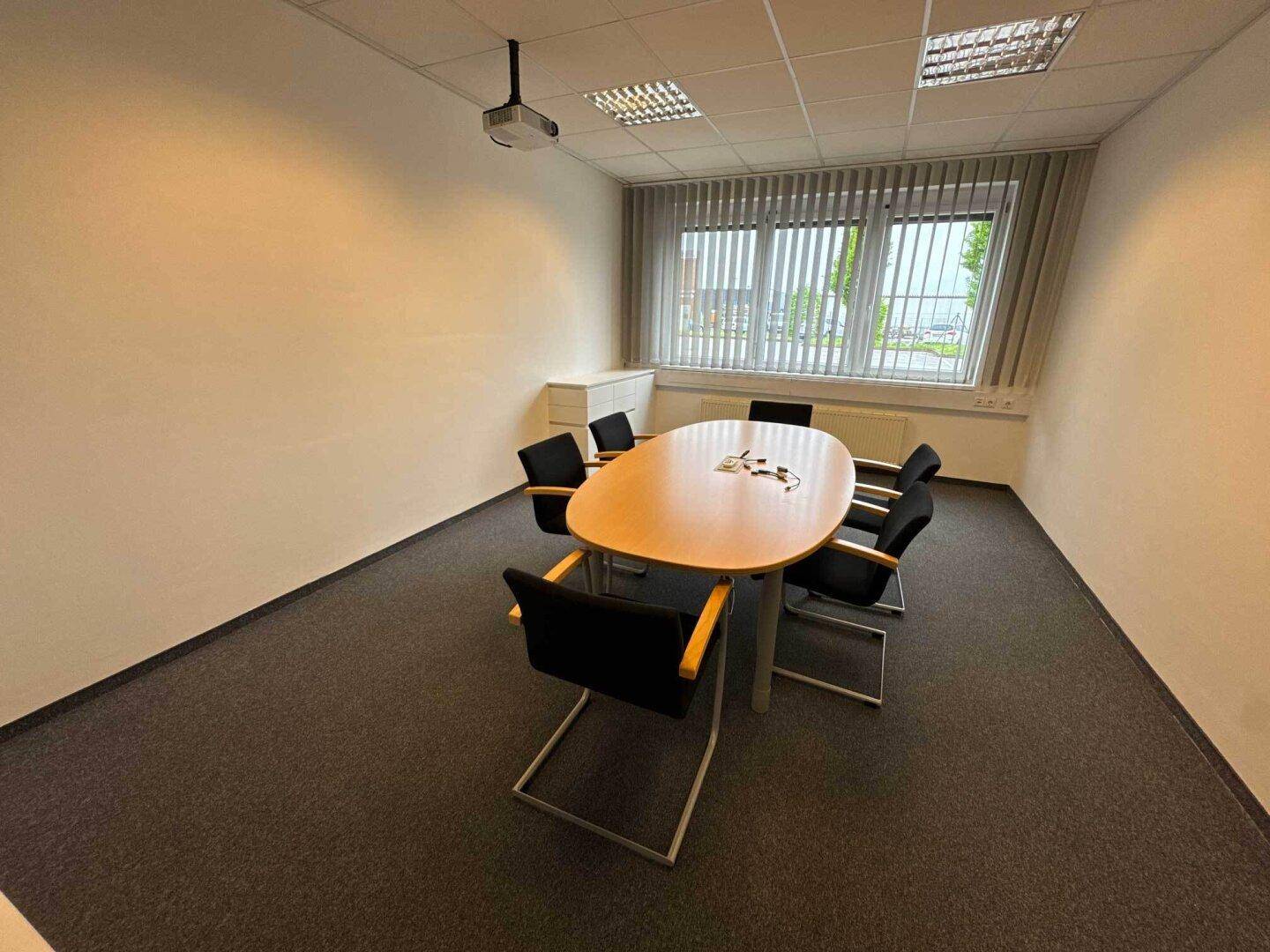 Konferenzraum mit ovalem Tisch, Teppichboden, Beamer und Jalousien an den Fenstern.