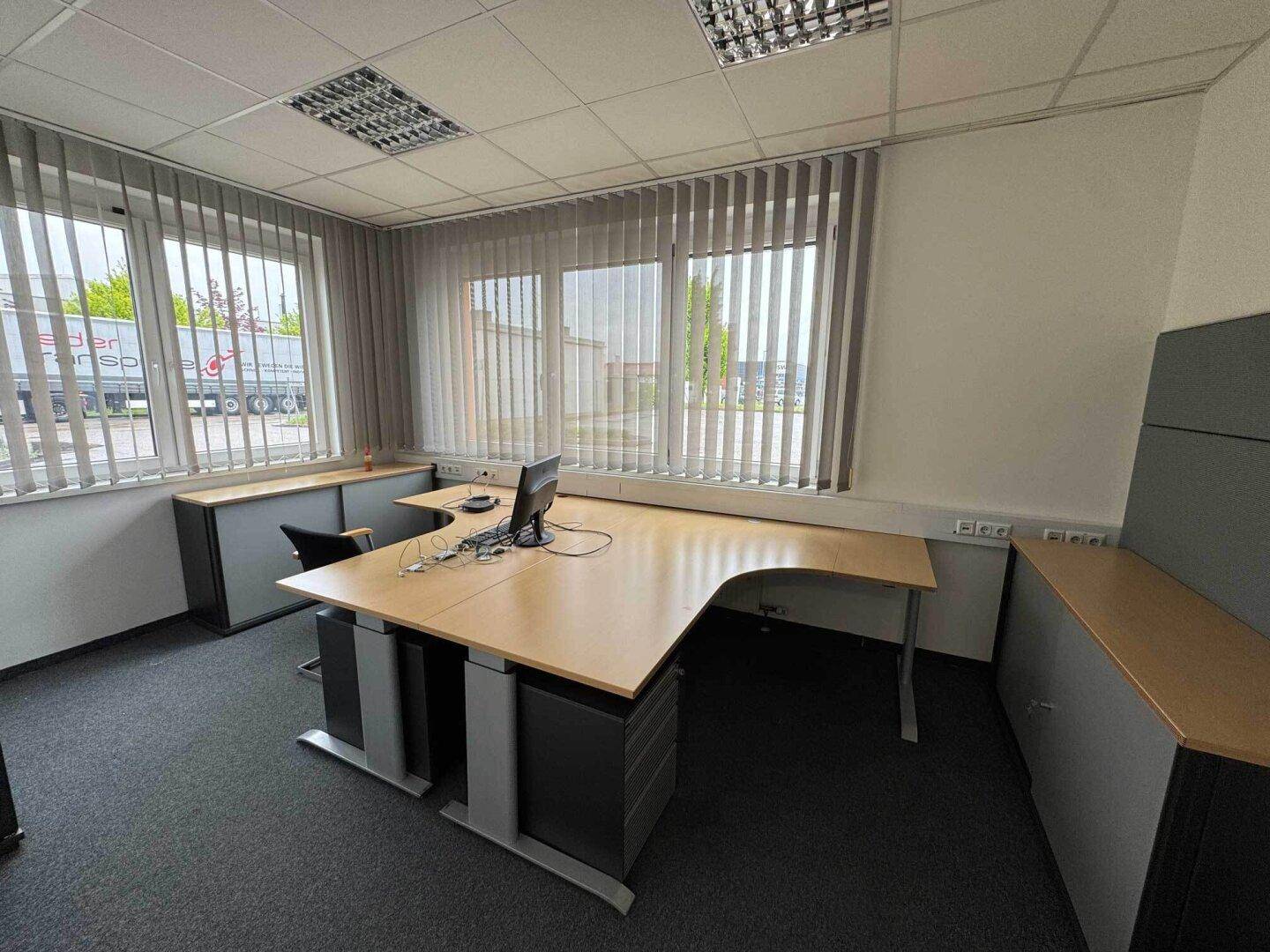 Helles Büro mit L-förmigem Schreibtisch, Teppichboden und Jalousien an den großen Fenstern.