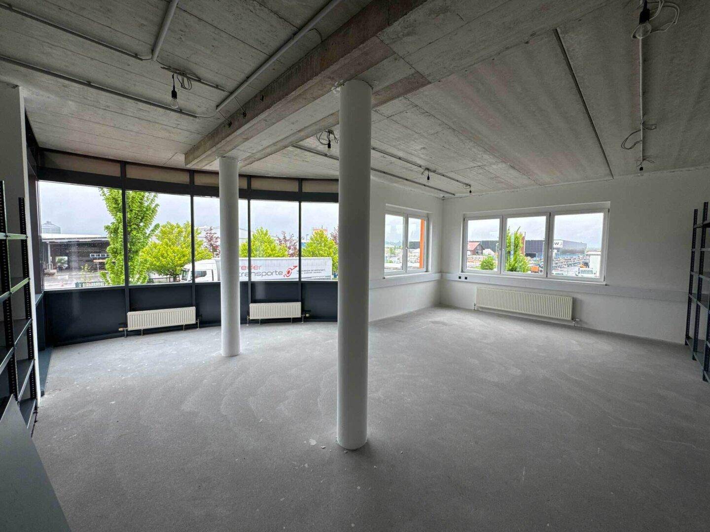 Großer, leerer Büroraum mit Betonboden, Säulen und bodentiefen Fenstern, die viel Licht hereinlassen.