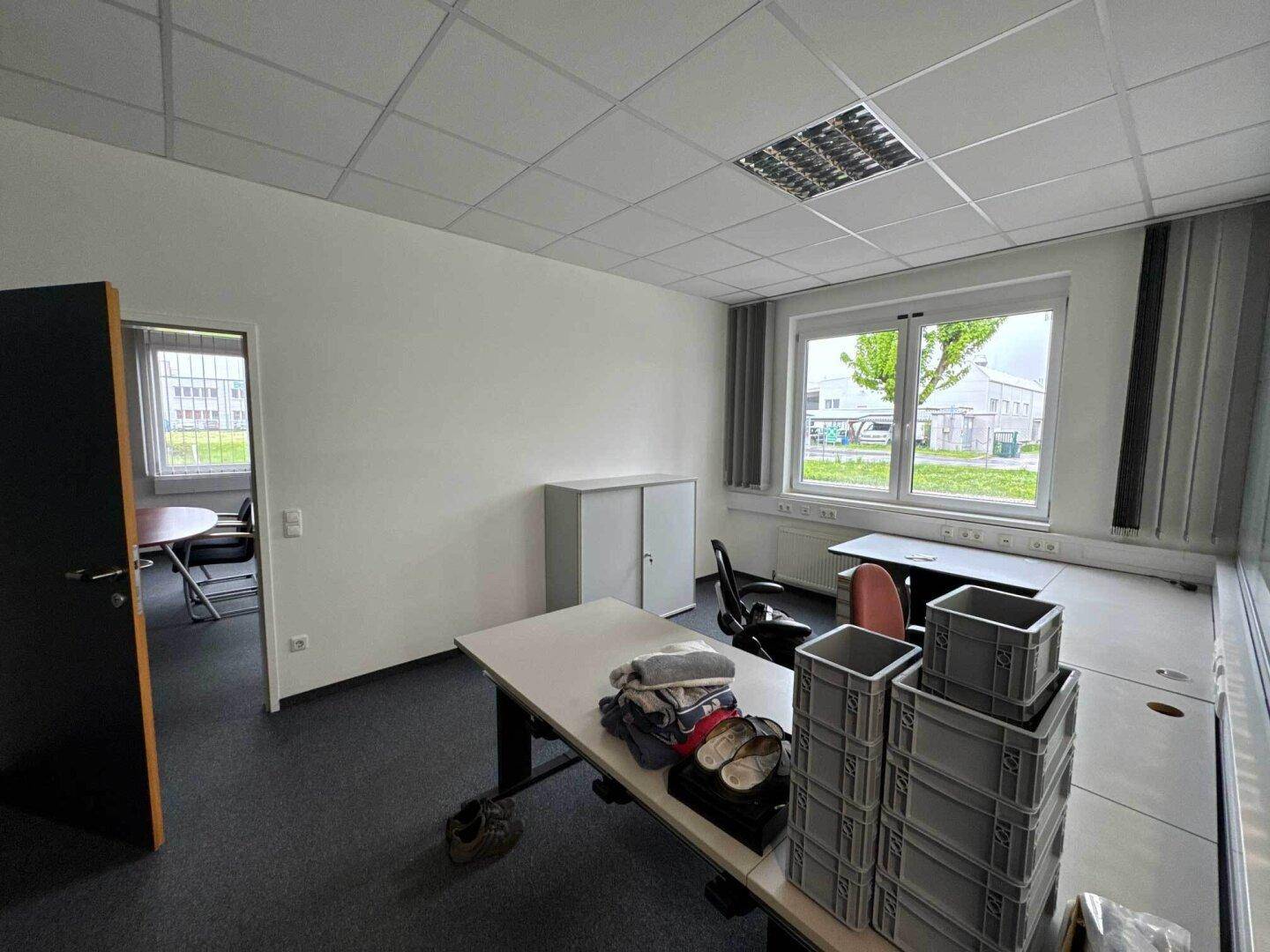 Büroraum mit Teppichboden, Schreibtischen und einem Fenster mit Blick auf das Außengelände.