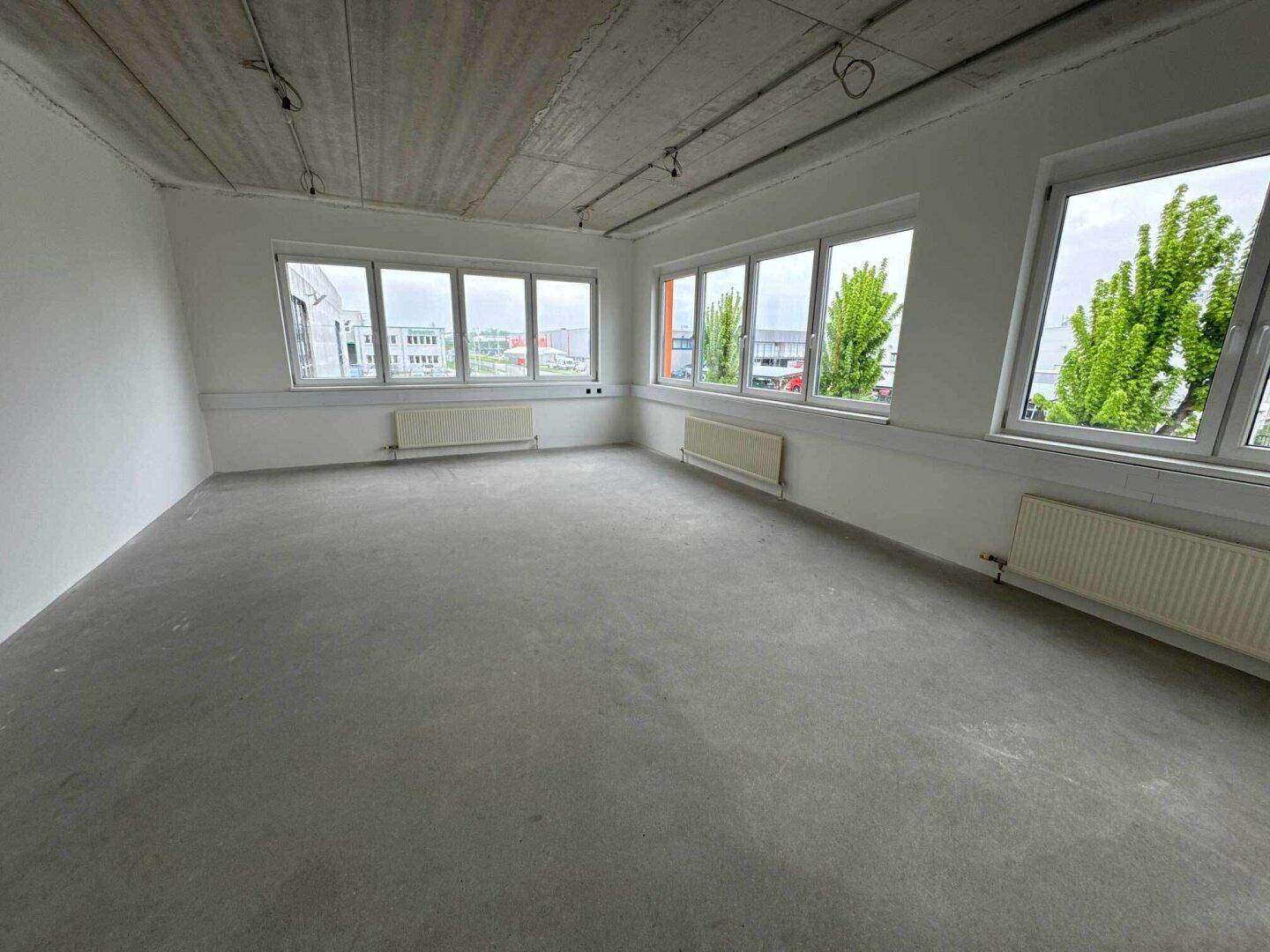 Heller Büroraum mit Betonboden, Heizkörpern und großen Fenstern, die eine weite Aussicht bieten.