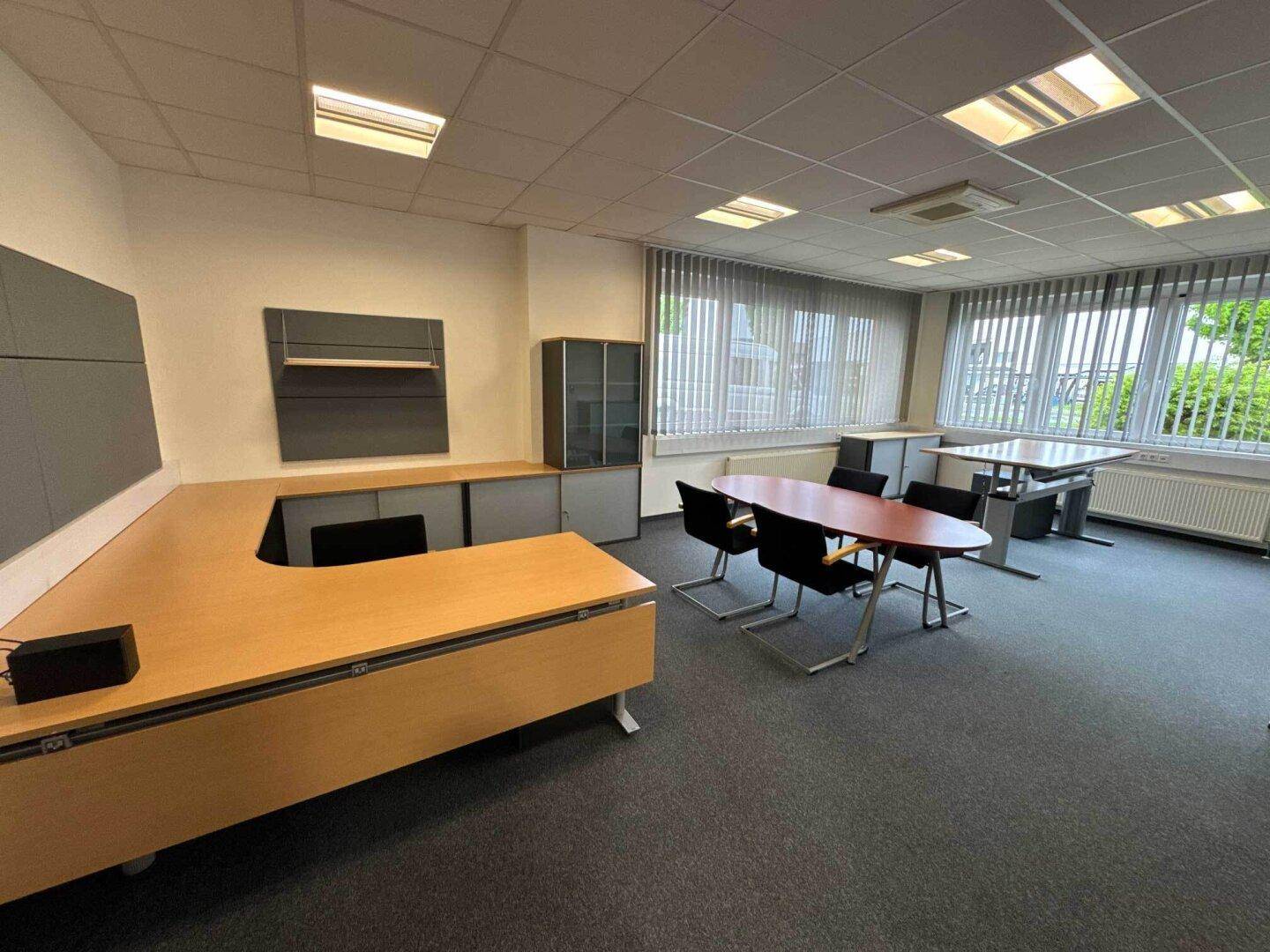 Geräumiges Büro mit mehreren Arbeitsplätzen, Teppichboden und großen Fenstern mit Jalousien.