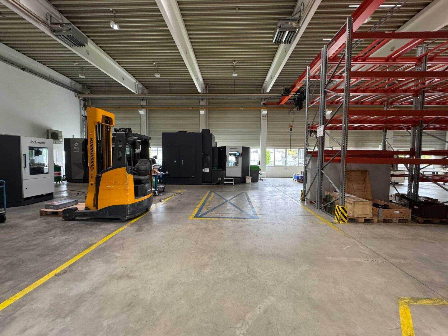 Weitläufige Lagerhalle mit Gabelstapler, Maschinen und großen Fenstern, die viel Tageslicht hereinlassen.