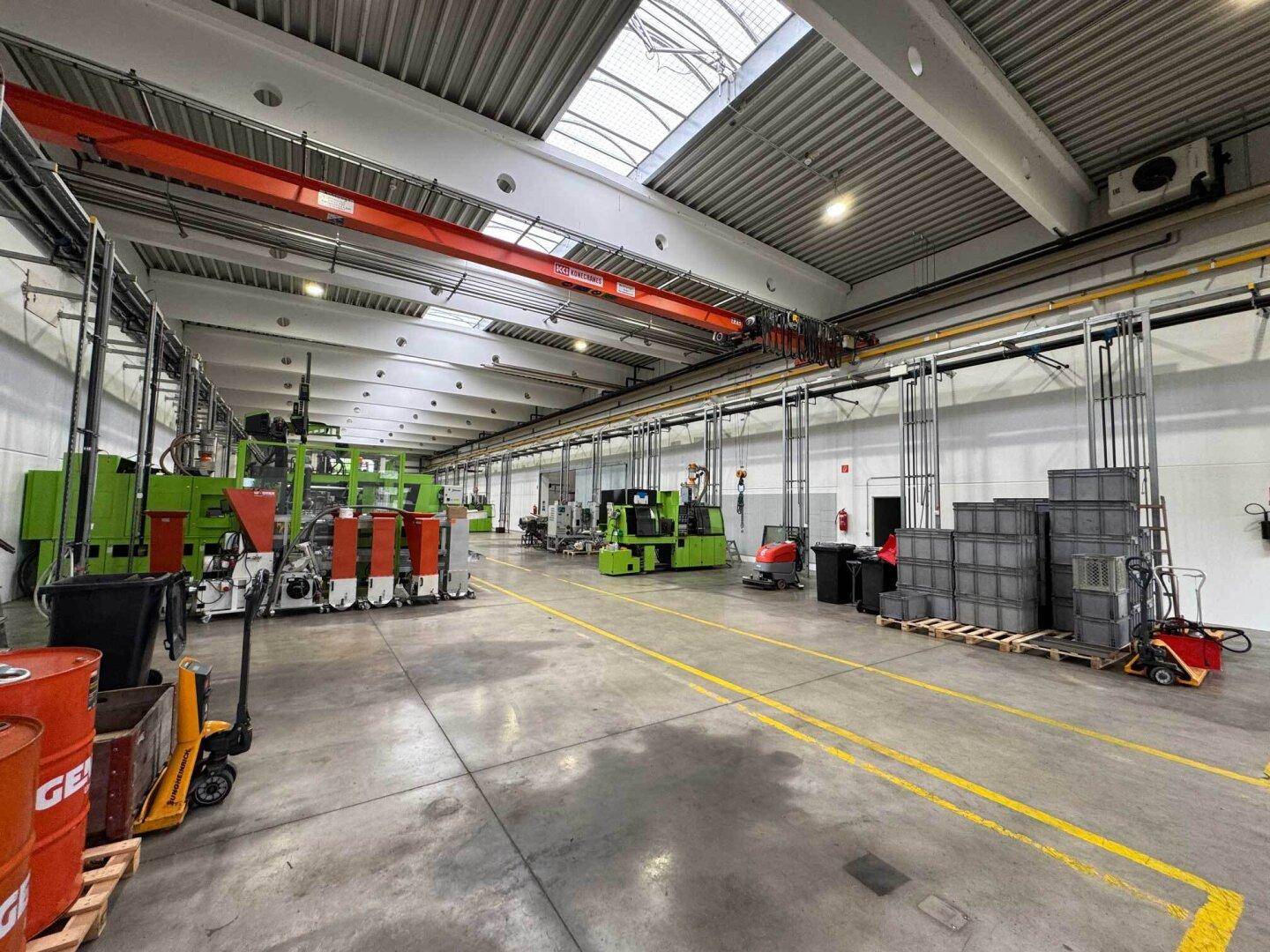 Lange Produktionshalle mit Kranbahn, Maschinen und Lagerbehältern, gut beleuchtet durch Oberlichter.