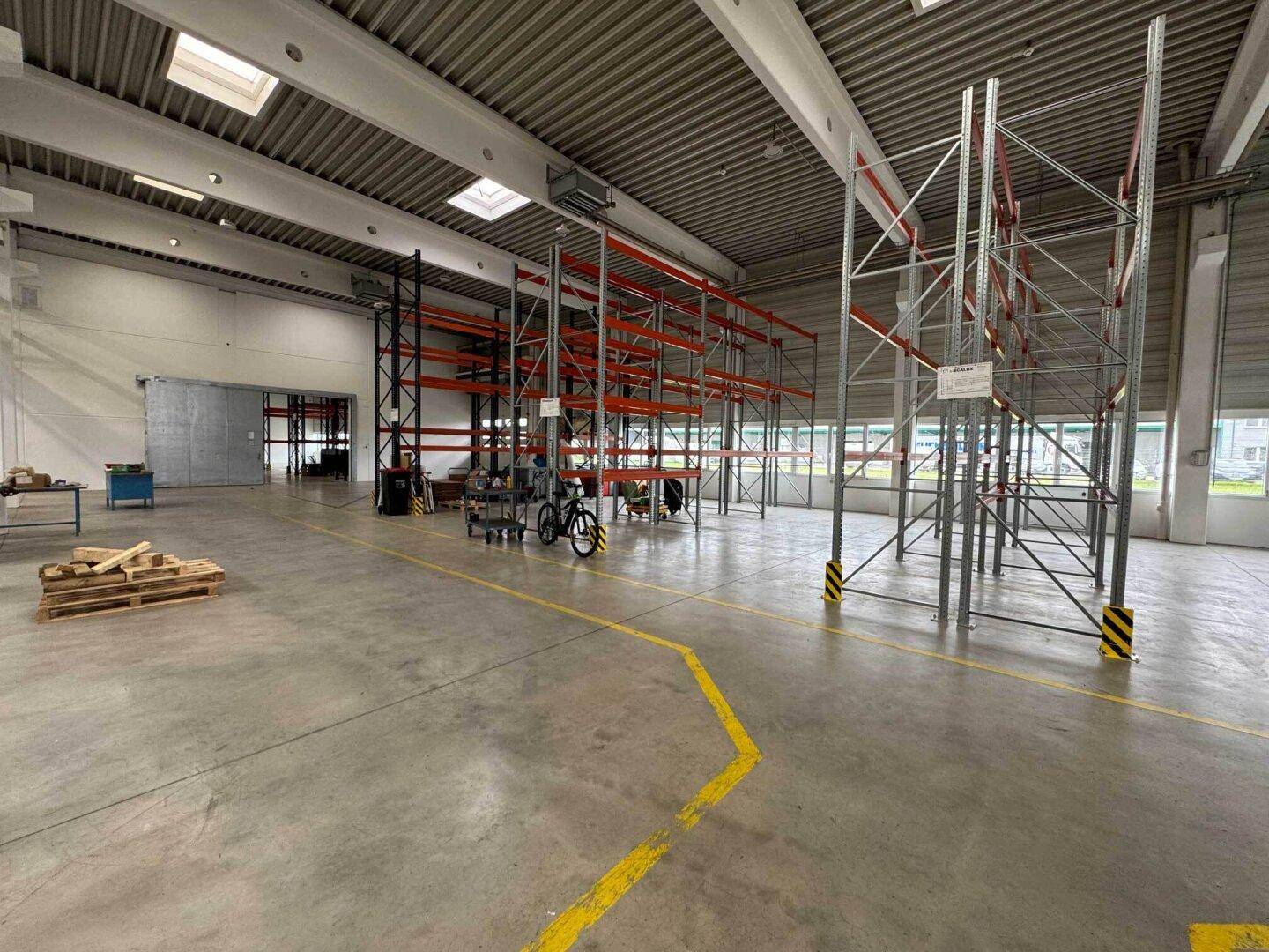 Geräumige Lagerhalle mit hohen Decken, leeren Regalsystemen und Betonboden mit gelben Markierungen.