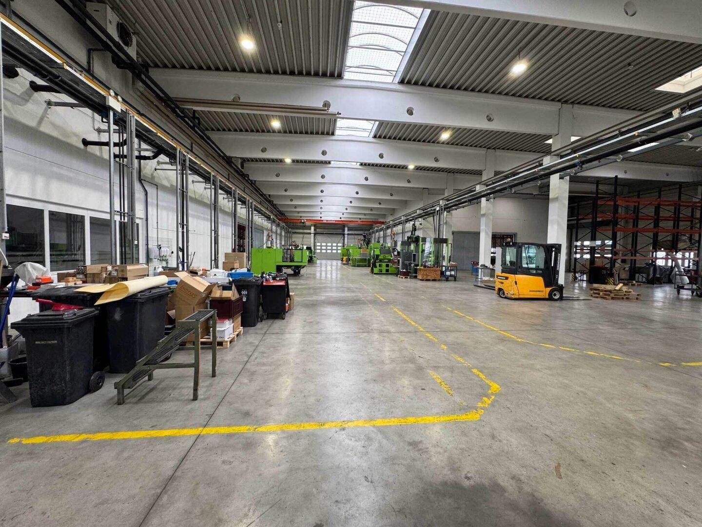 Übersicht über die Produktionshalle mit Maschinen, Gabelstapler und Mülltonnen, mit Zugangstüren im Hintergrund.