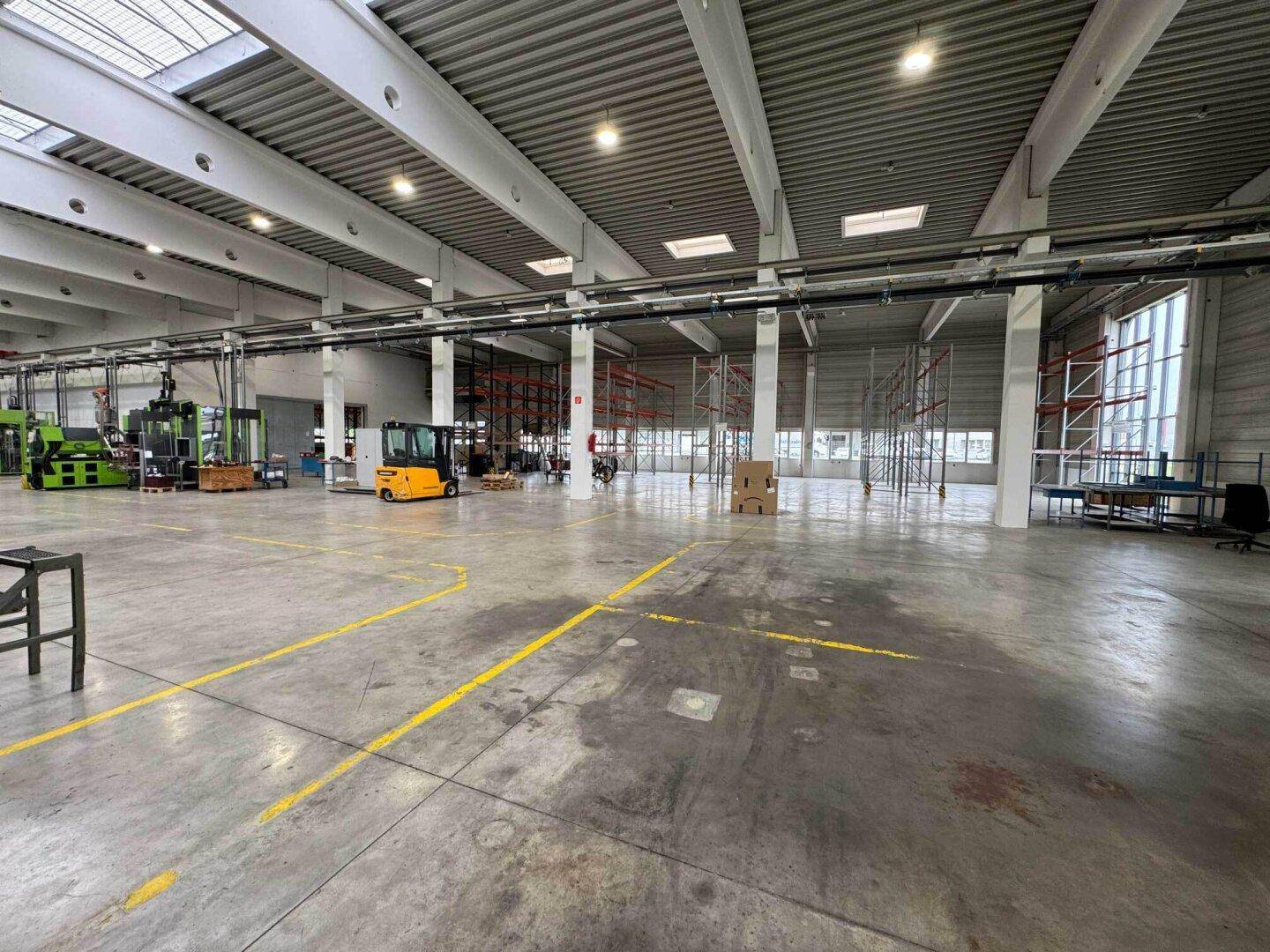 Leere Produktionsfläche in einer Halle mit hohen Decken, Oberlichtern und Regalsystemen im Hintergrund.