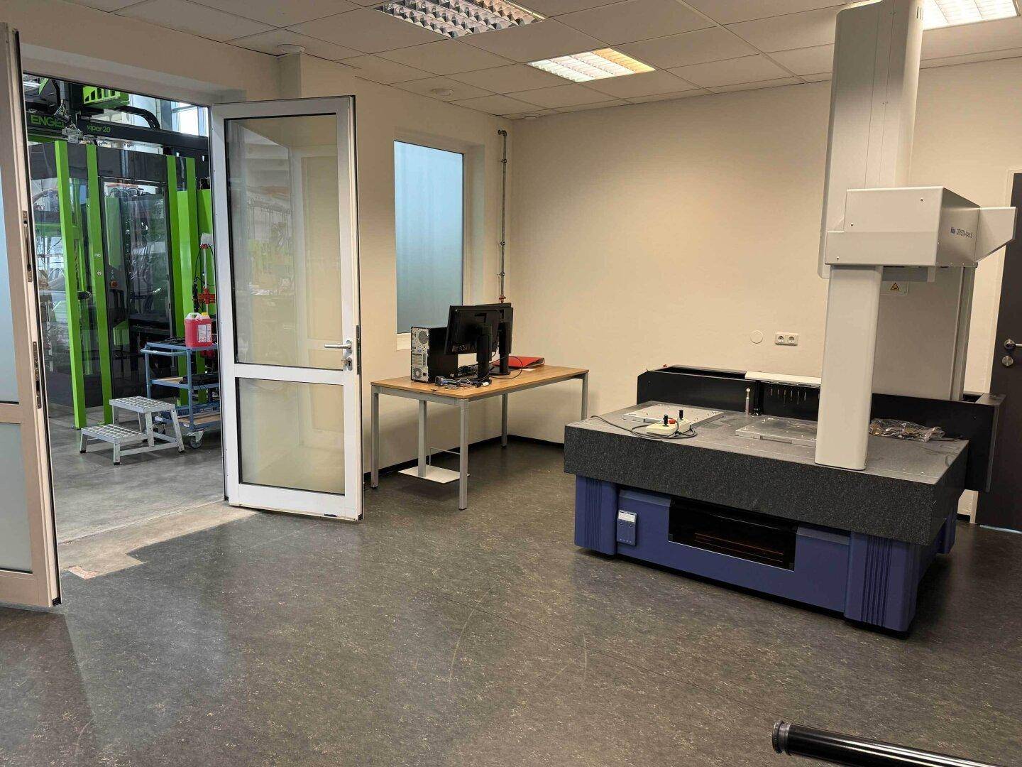 Büro- oder Laborraum mit Präzisionsmessmaschine, Schreibtisch und Blick in einen Produktionsbereich.