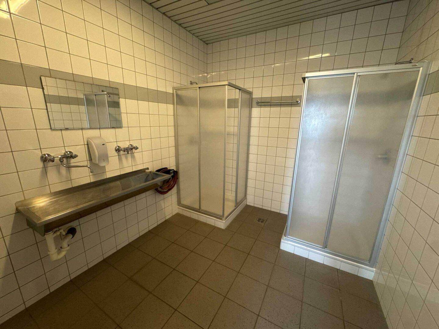 Umkleide- und Duschraum mit mehreren Duschkabinen, langem Waschbecken und gefliesten Wänden.