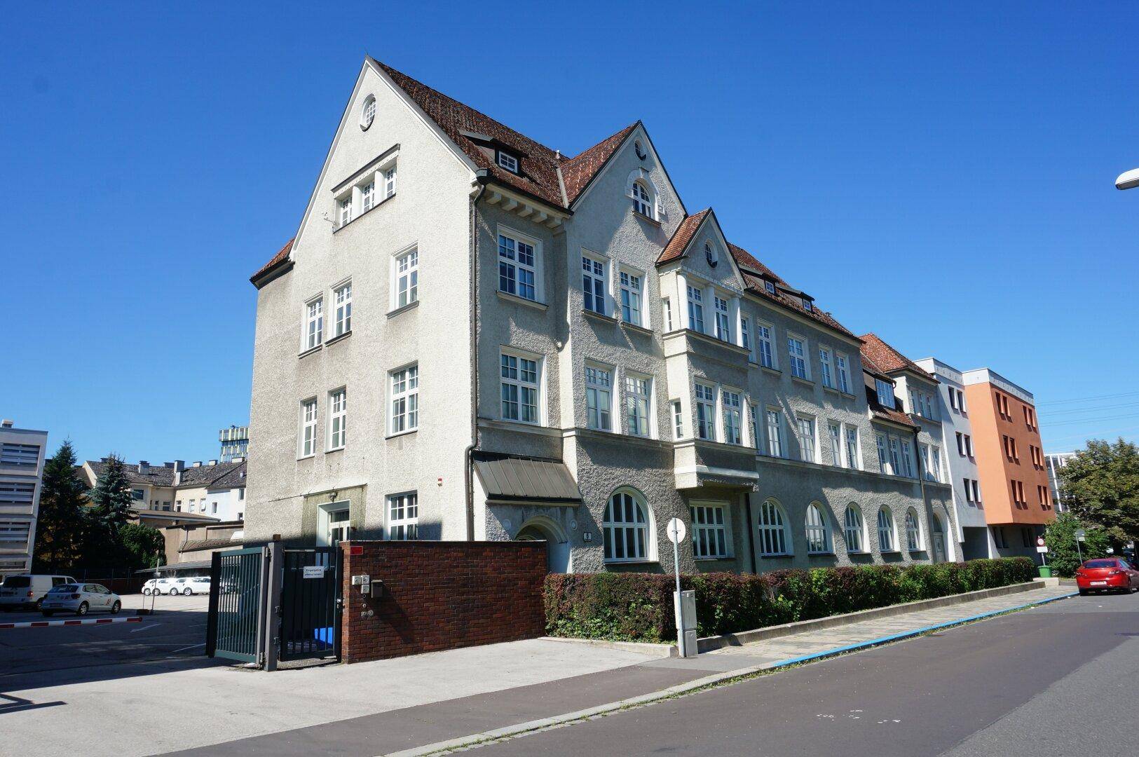 Historisches Gebäude mit heller Fassade, vielen Fenstern und einem roten Ziegeldach, umgeben von einer Straße und Parkplätzen.