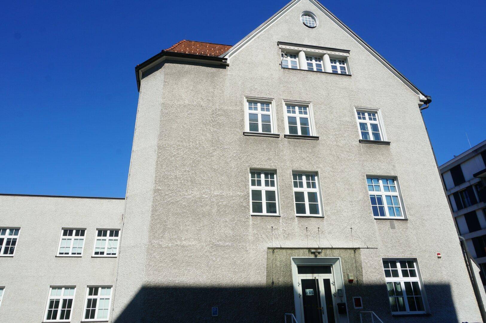 Detailansicht der hellen Fassade eines Altbaus mit vielen weißen Fenstern und einem markanten Giebel unter klarem Himmel.