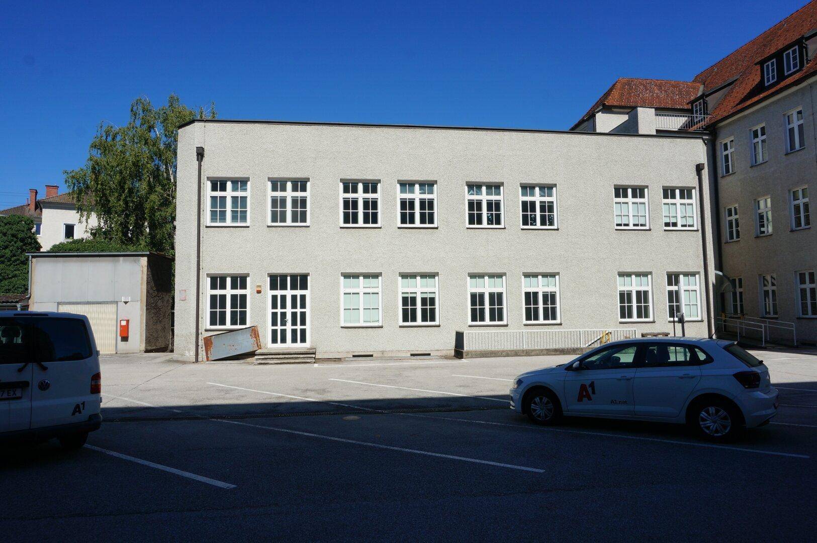 Rückansicht eines Gebäudes mit heller Fassade, vielen Fenstern und einem angrenzenden Parkplatzbereich.