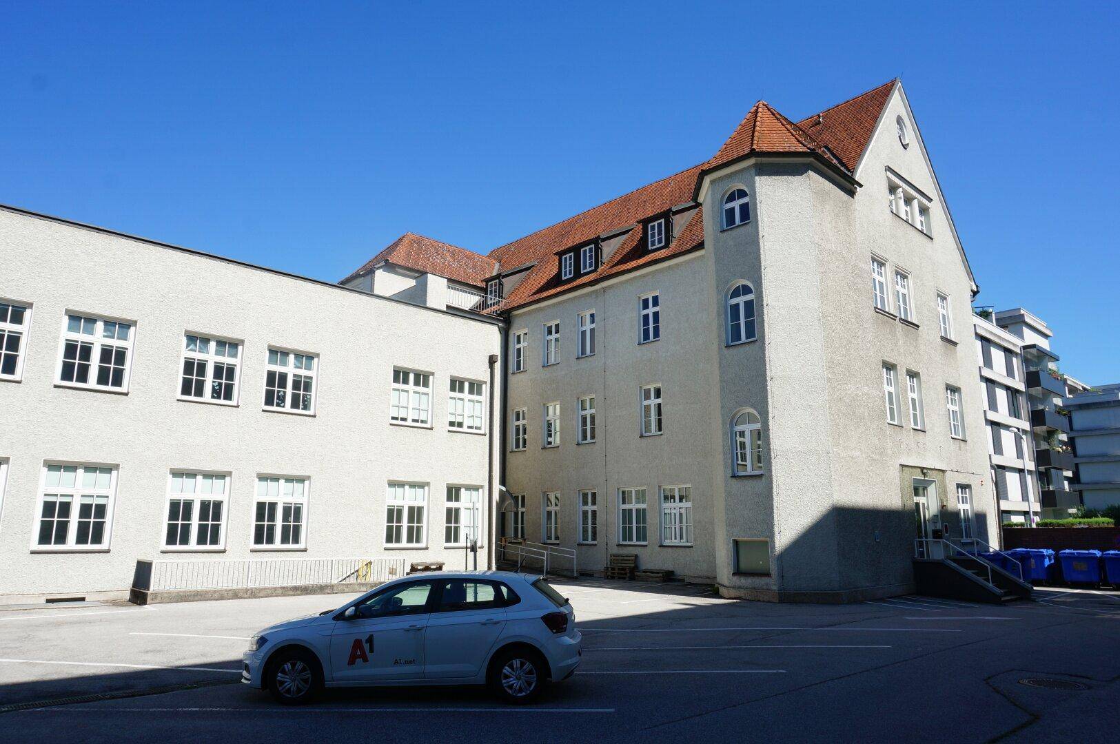 Ansicht des Gebäudes mit heller Fassade, roten Ziegeldächern und einem angrenzenden Parkplatz unter blauem Himmel.
