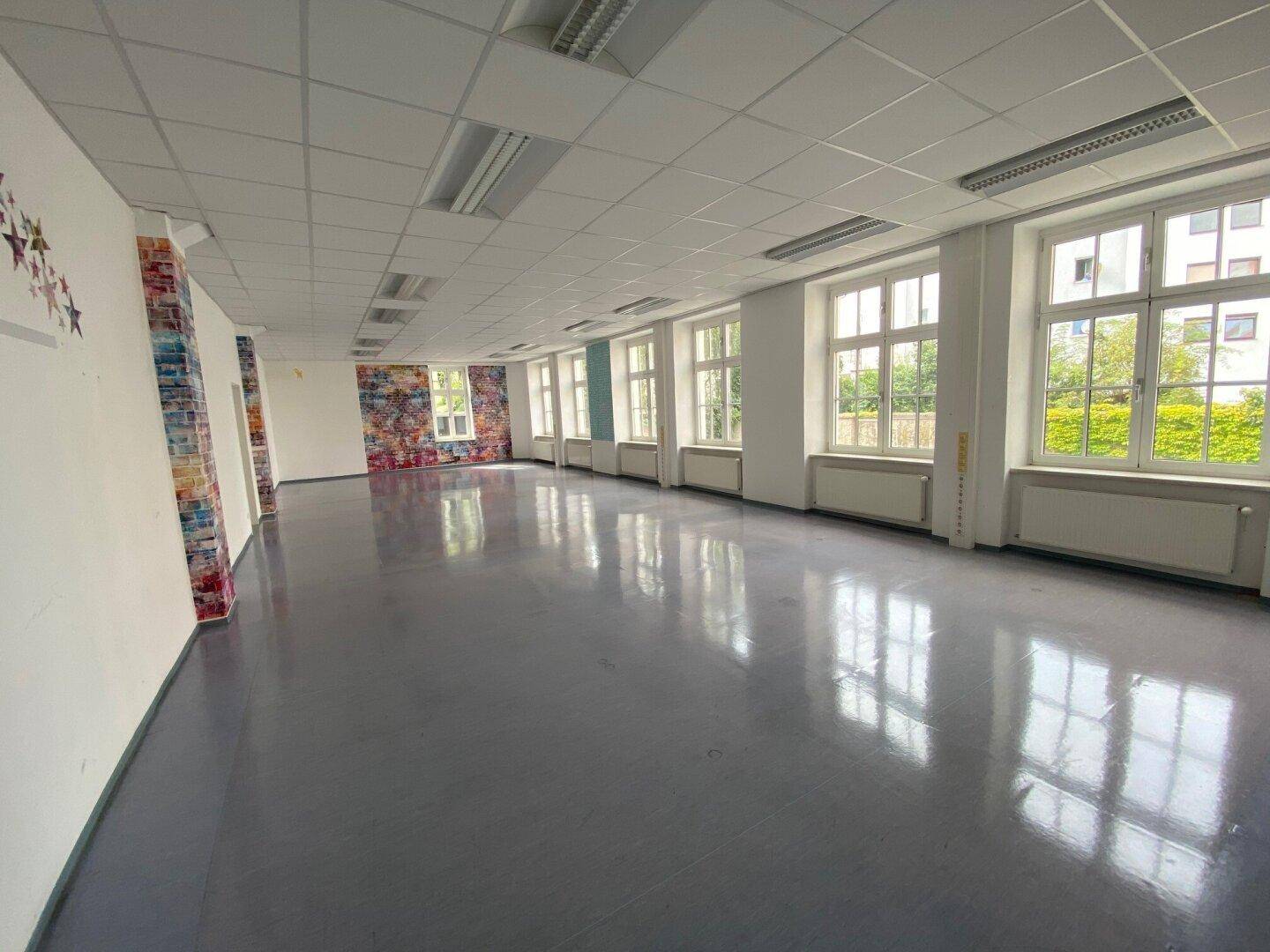 Großer, leerer Raum mit glänzendem grauen Boden, vielen Fenstern und einer abgehängten Decke.