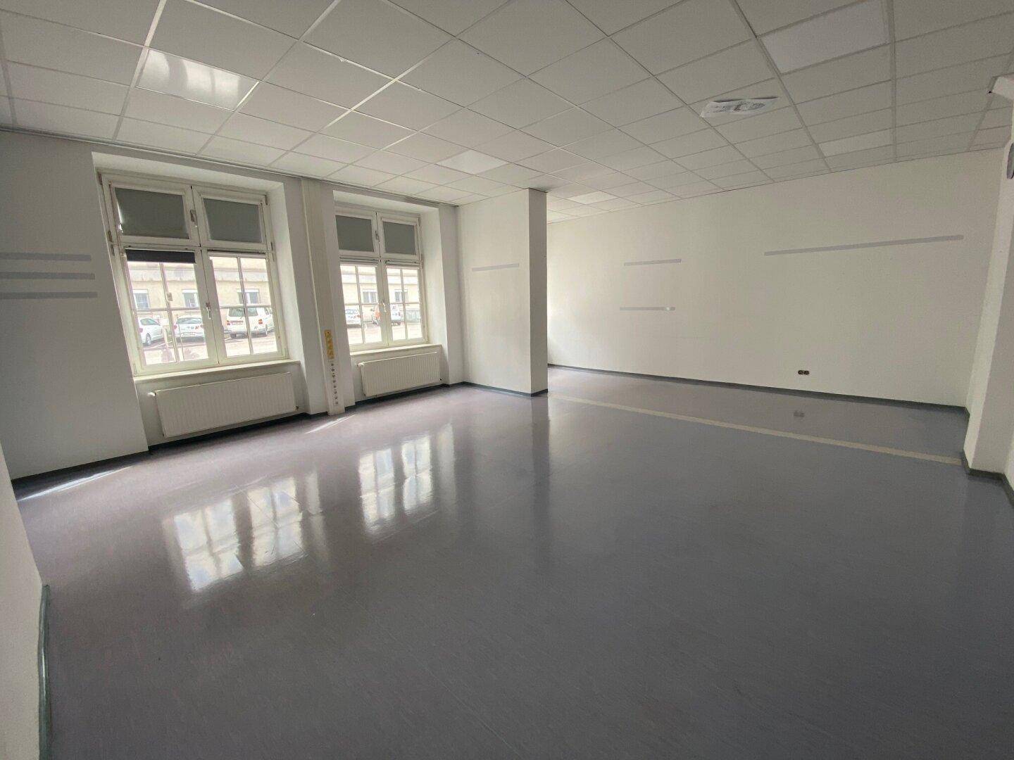 Heller, leerer Raum mit glänzendem grauen Boden, großen Fenstern und einer schlichten weißen Decke.