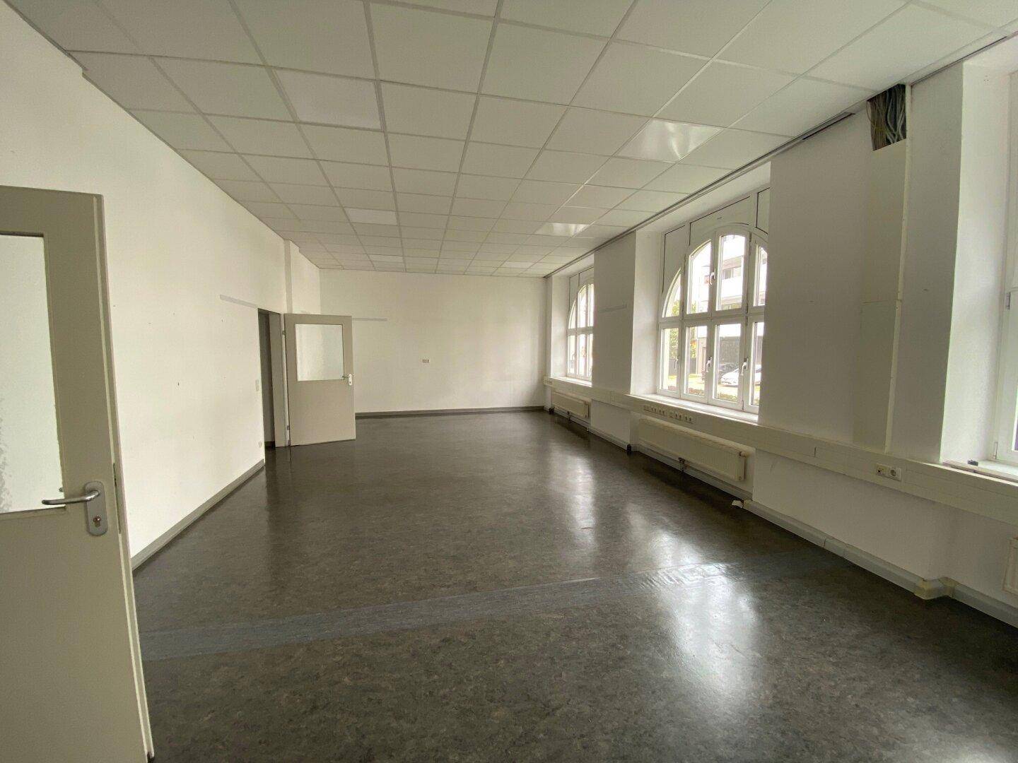 Geräumiger, leerer Raum mit glänzendem dunklem Boden, hohen Fenstern und einer weißen Decke.