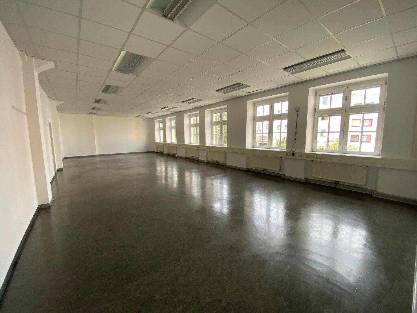 Großer, heller Raum mit glänzendem dunklem Boden, vielen Fenstern und einer abgehängten Decke.