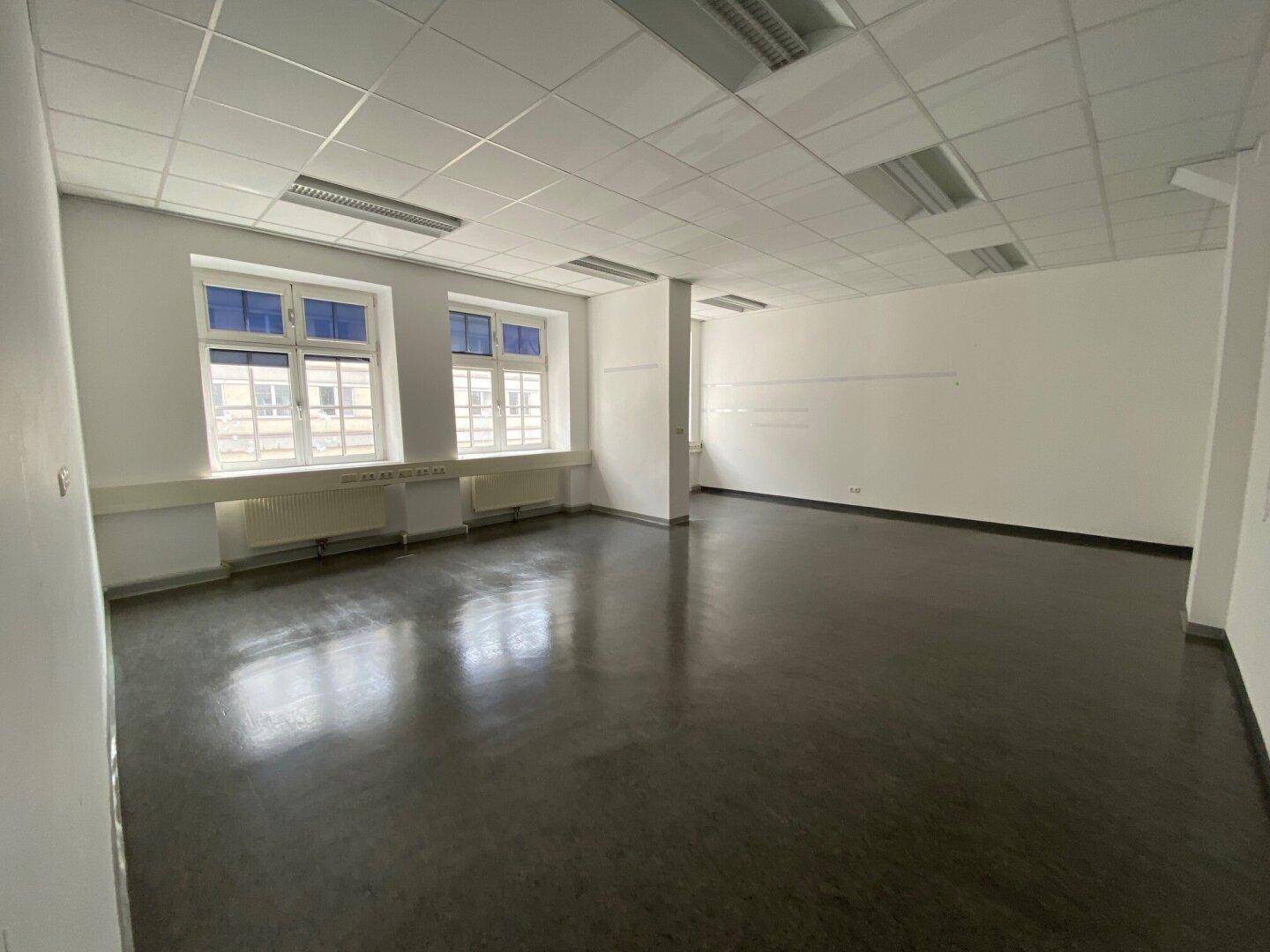 Heller, leerer Raum mit glänzendem dunklem Boden, großen Fenstern und einer weißen Decke.