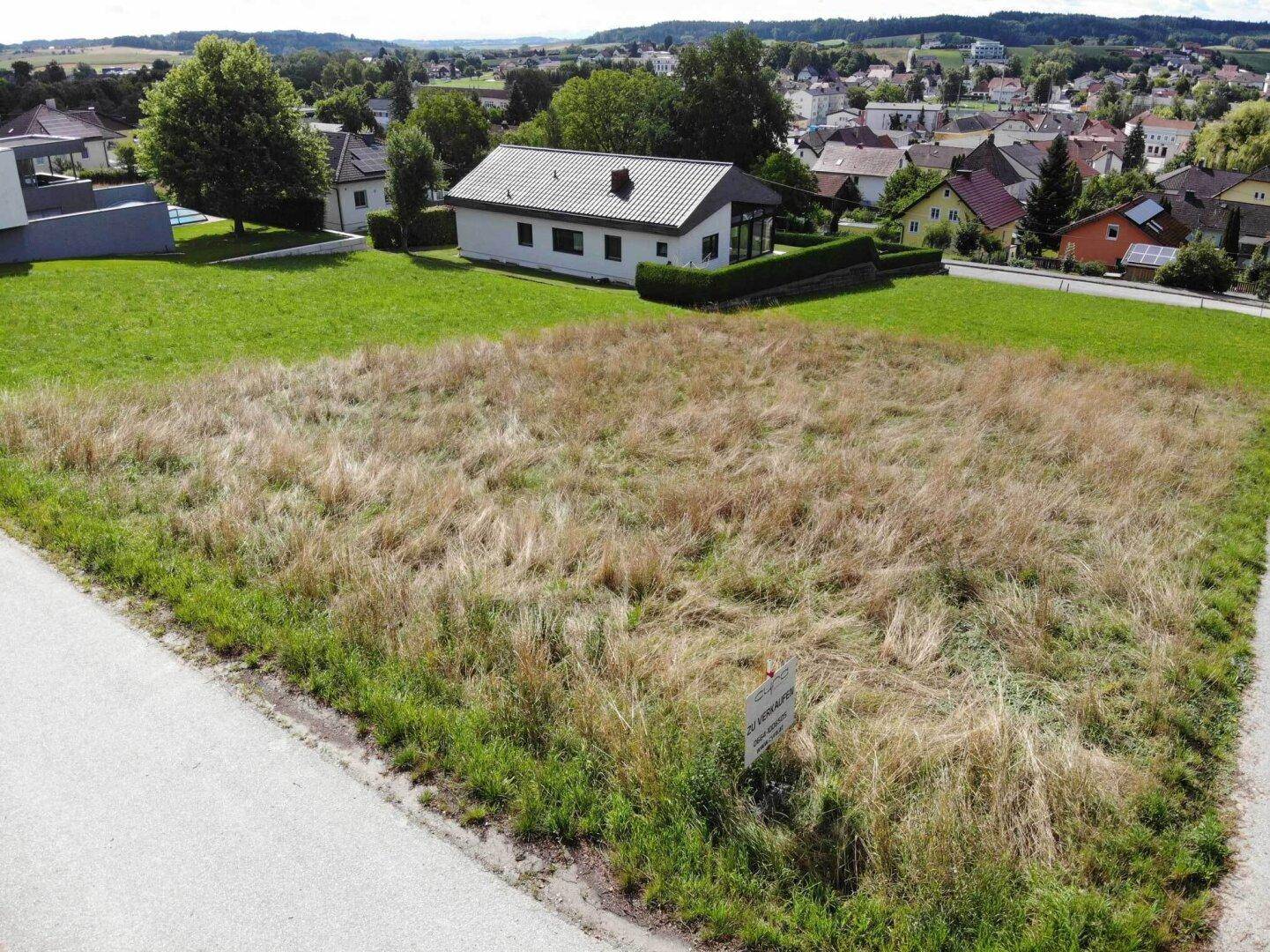 Baugrundstück aus Bodennähe mit Verkaufsanzeige, umgeben von grüner Landschaft und bestehenden Wohnhäusern.