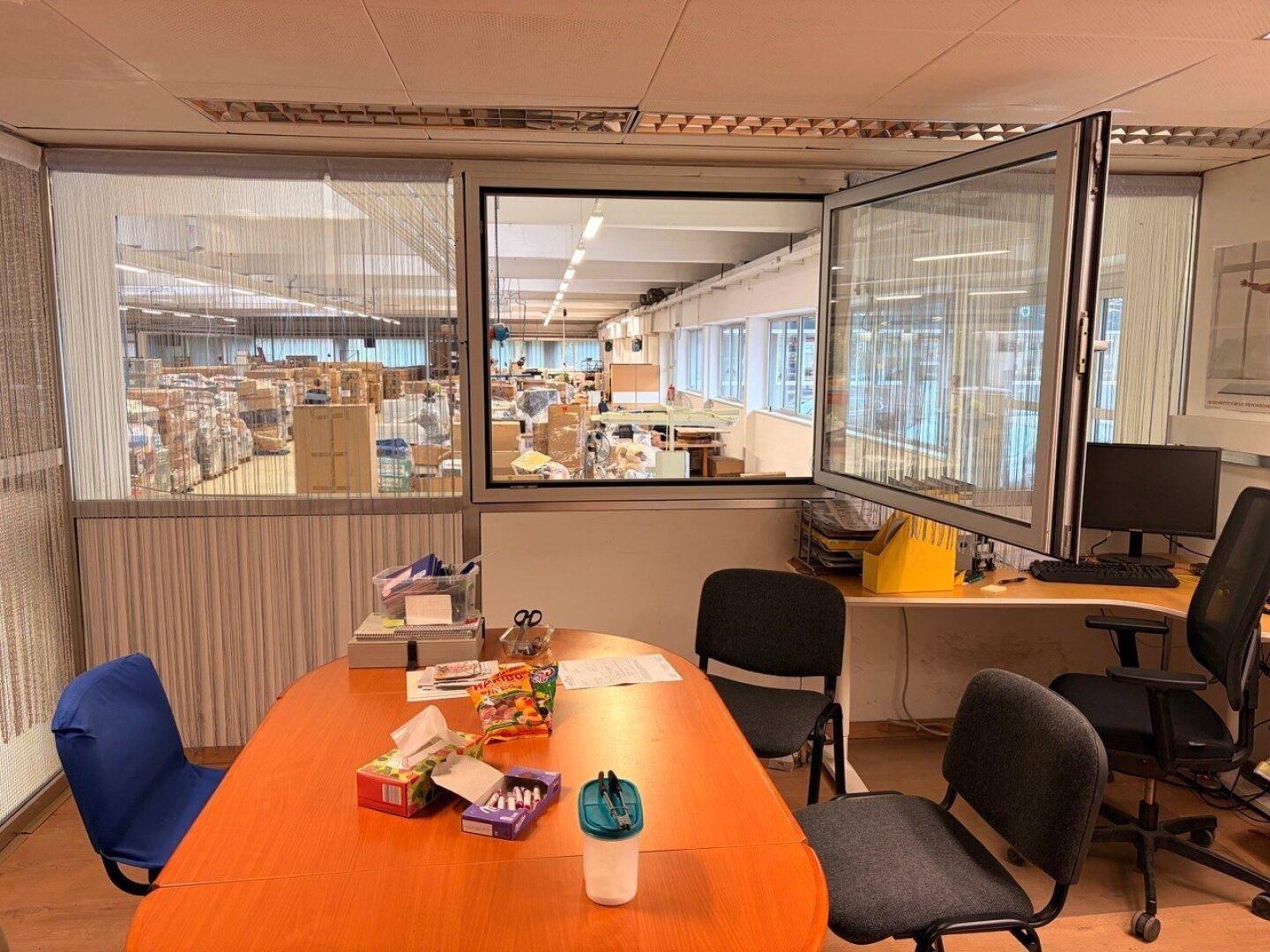 Büro mit Holztisch und Blick durch ein Fenster in die angrenzende Produktionshalle.