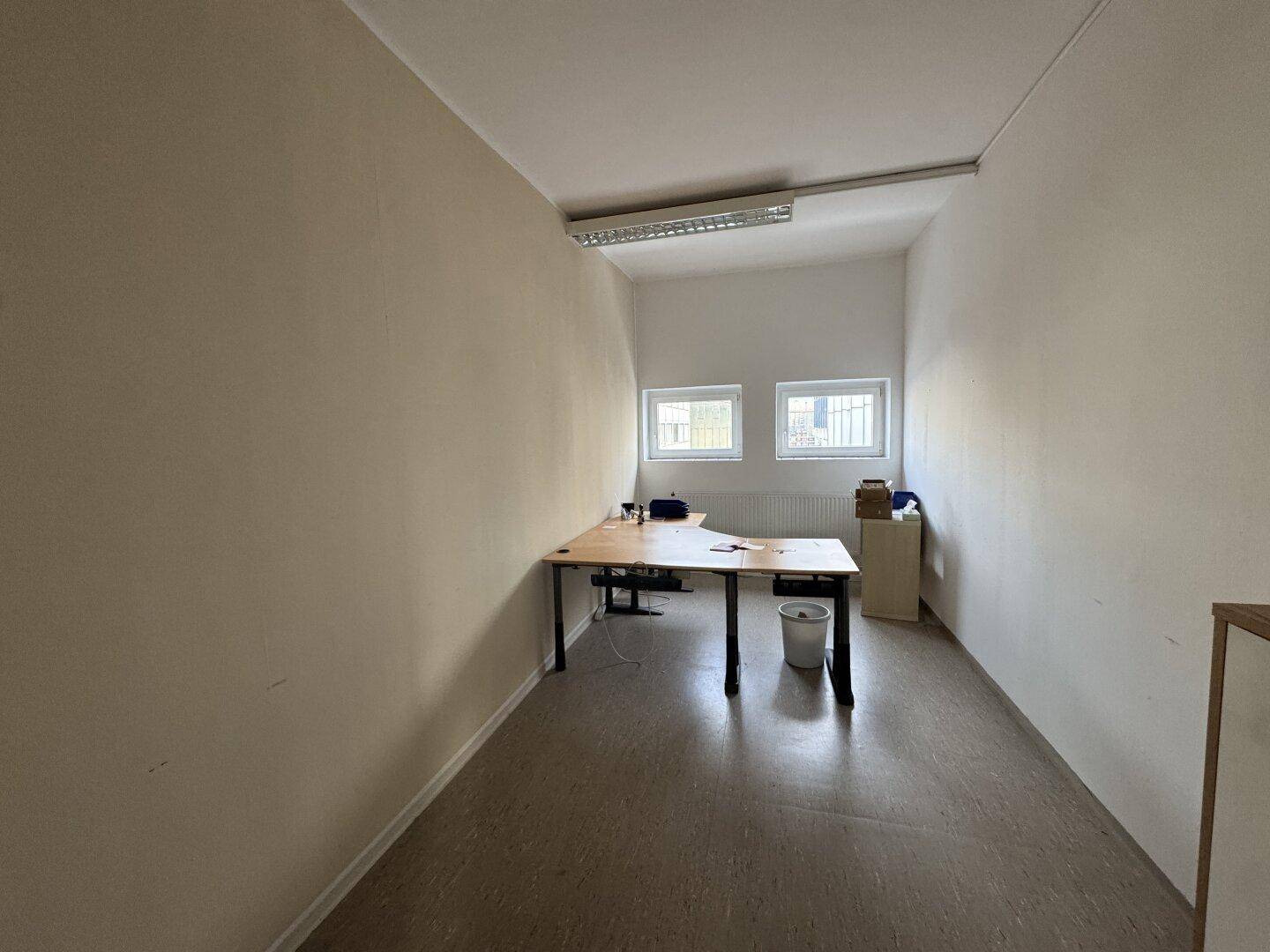 Funktionales Büro mit zwei Fenstern, einem Schreibtisch und einfacher Einrichtung.