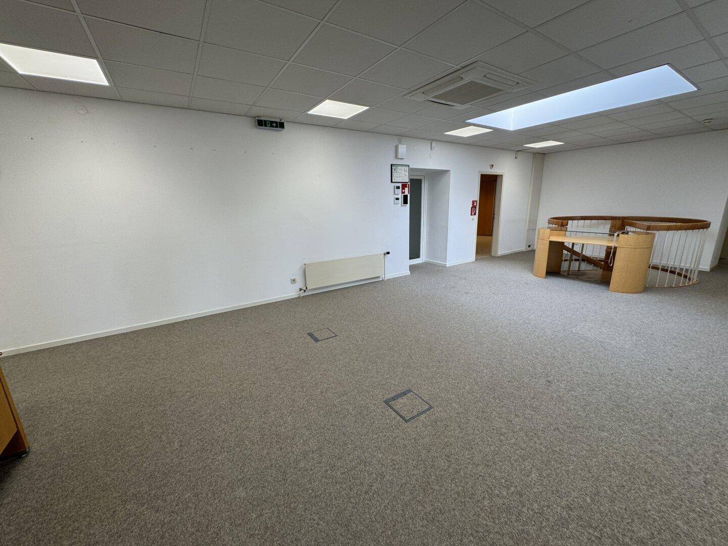 Großer offener Bürobereich mit Teppichboden, Deckenbeleuchtung und einer Wendeltreppe, ideal für flexible Arbeitsplatzgestaltung.
