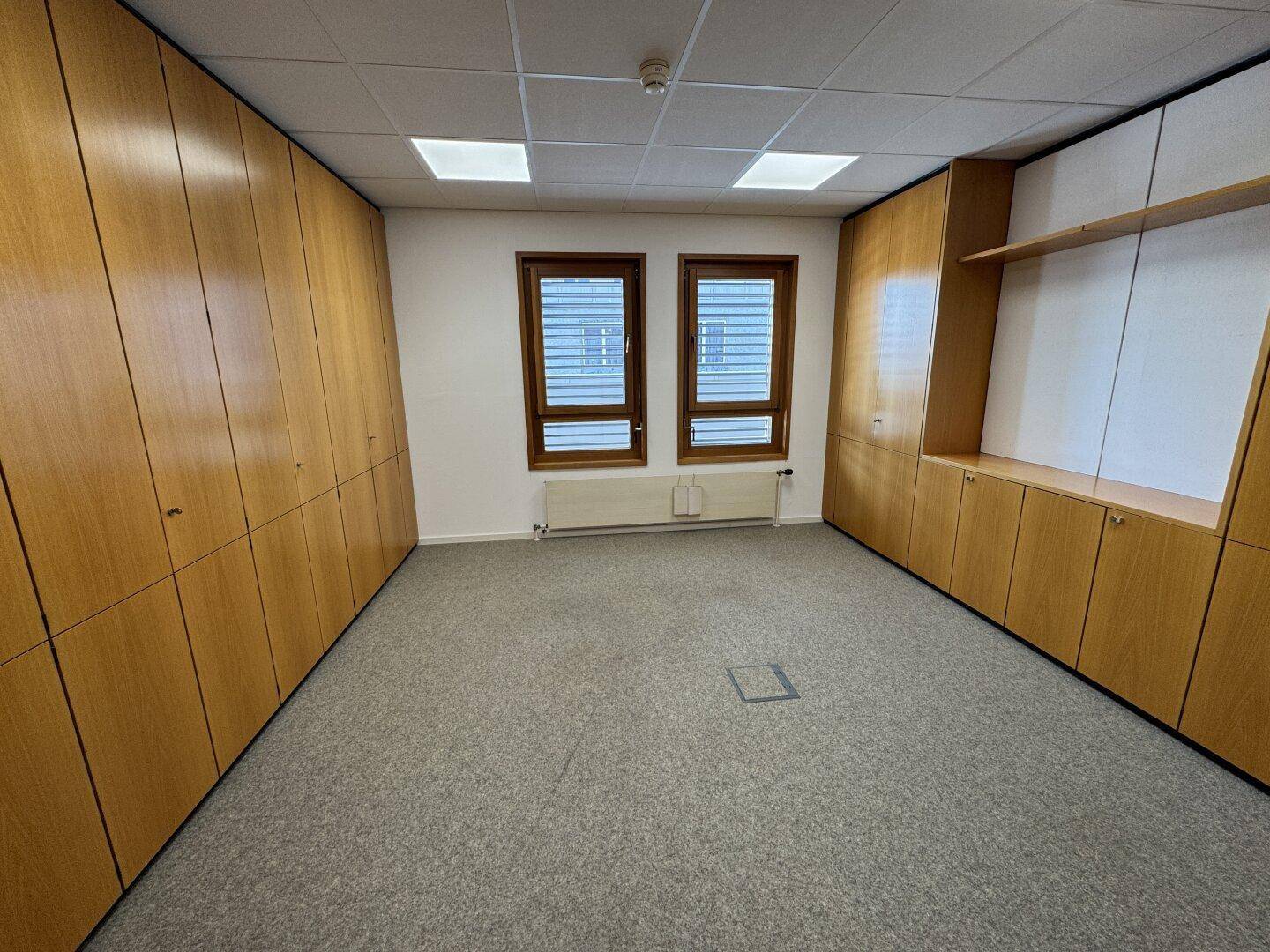 Leeres Büro mit Teppichboden, holzvertäfelten Wänden und integrierten Schränken, beleuchtet durch Deckenleuchten und Fenster.