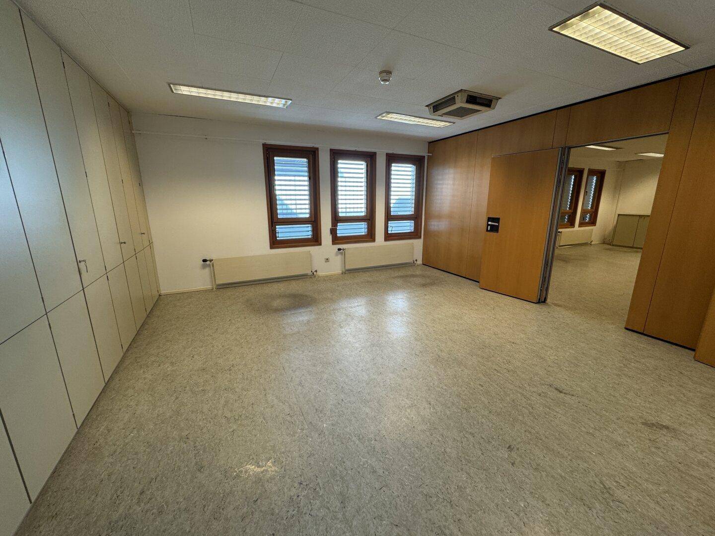 Geräumiges Büro mit Vinylboden, drei Fenstern mit Jalousien und Deckenbeleuchtung, bereit zur Einrichtung.