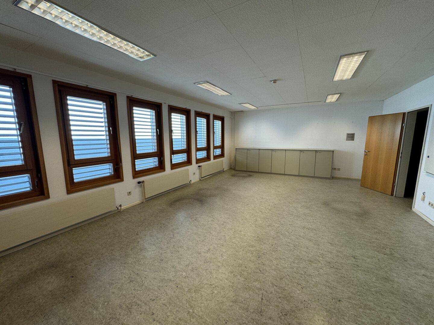 Helles Büro mit sechs Fenstern, Vinylboden und niedrigen Schränken entlang der Wand, ideal für Arbeitsbereiche.
